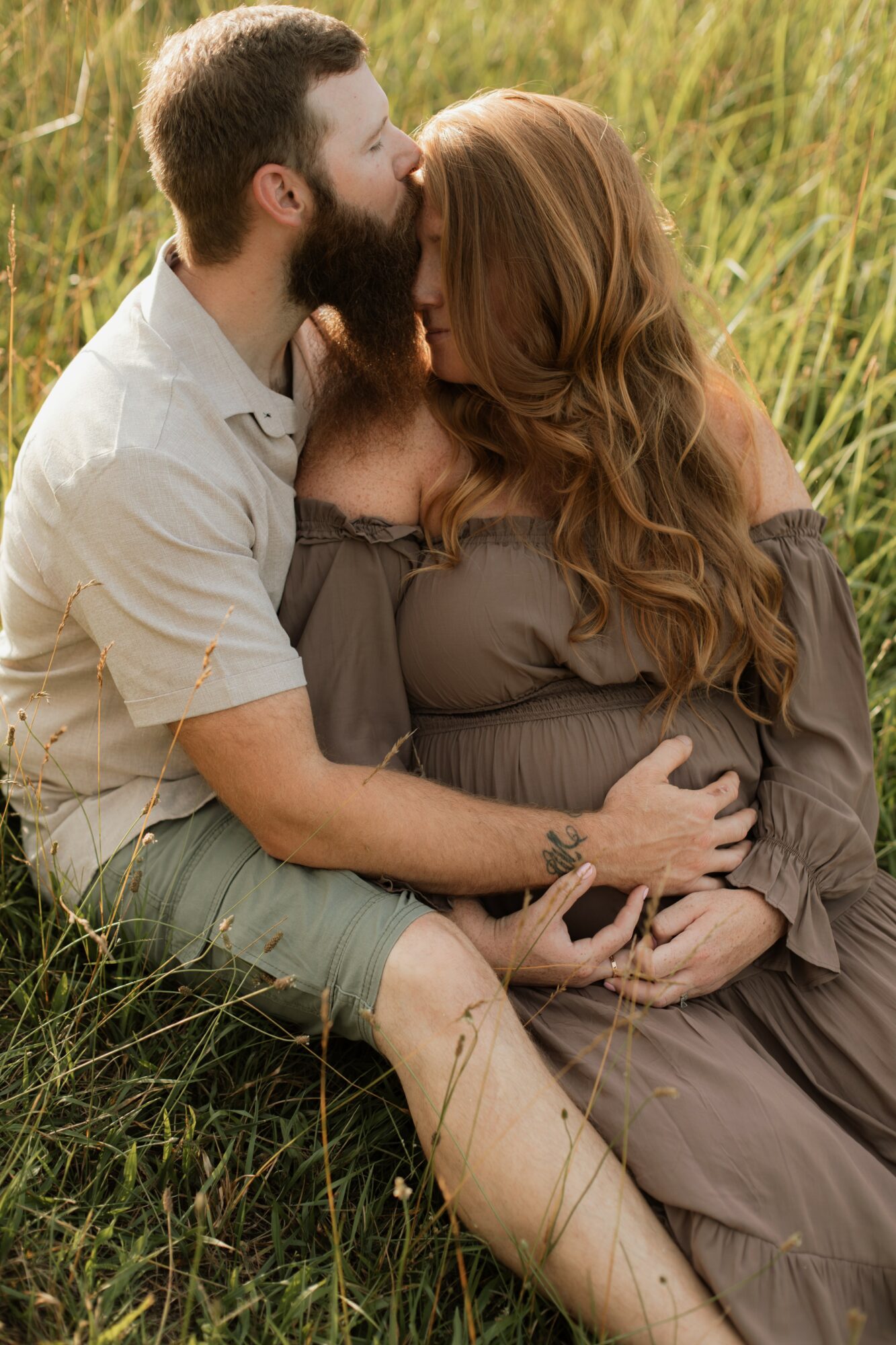 A couple sitting in a grassy field, the man with a beard and the woman with long red hair, embracing each other.