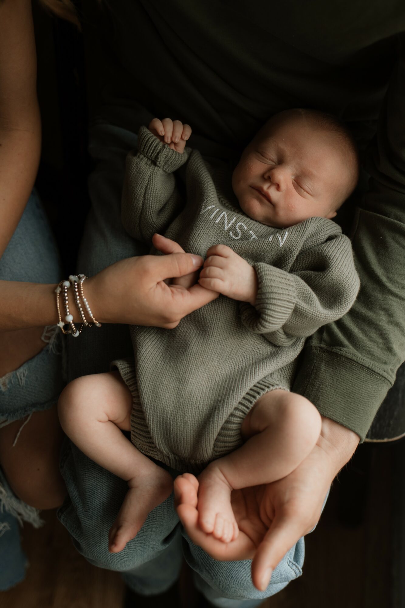 Baby sleeping in person's arms, wearing a gray sweater, with hand gently touching the baby's hand.