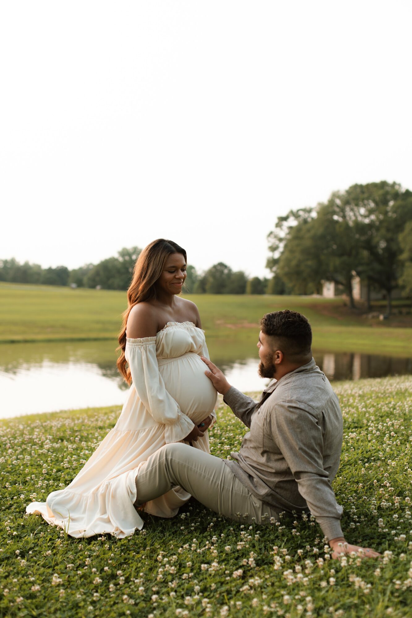Pregnant woman in a white dress sitting on grass, holding hands with a man sitting beside her outdoors near a lake.