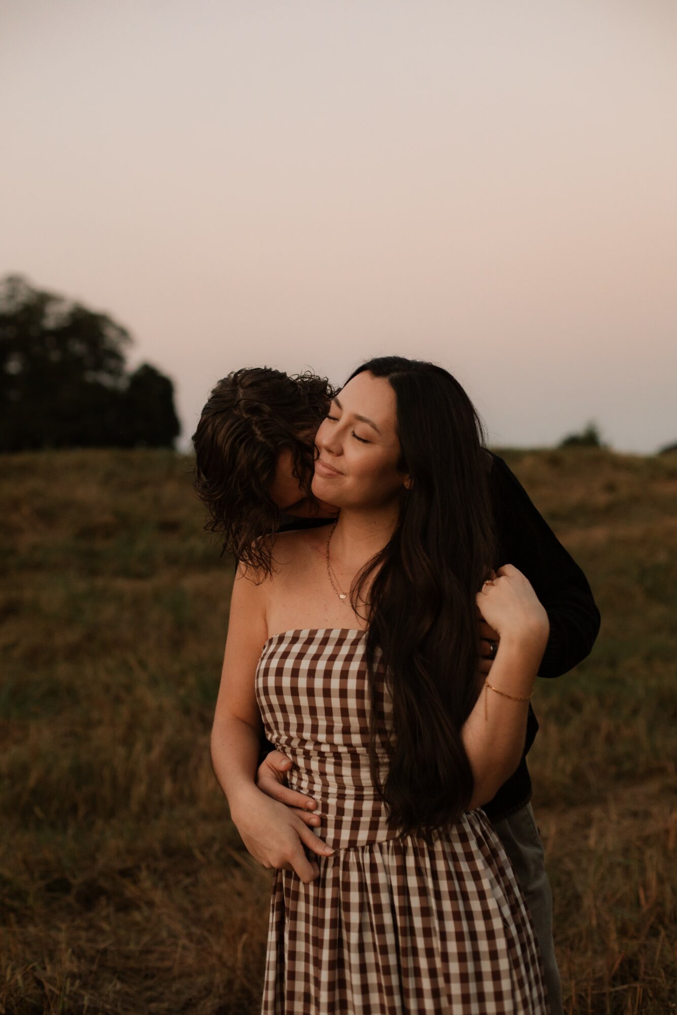 Two women embrace outdoors during sunset, one with long dark hair and the other with curly hair, in a grassy field.