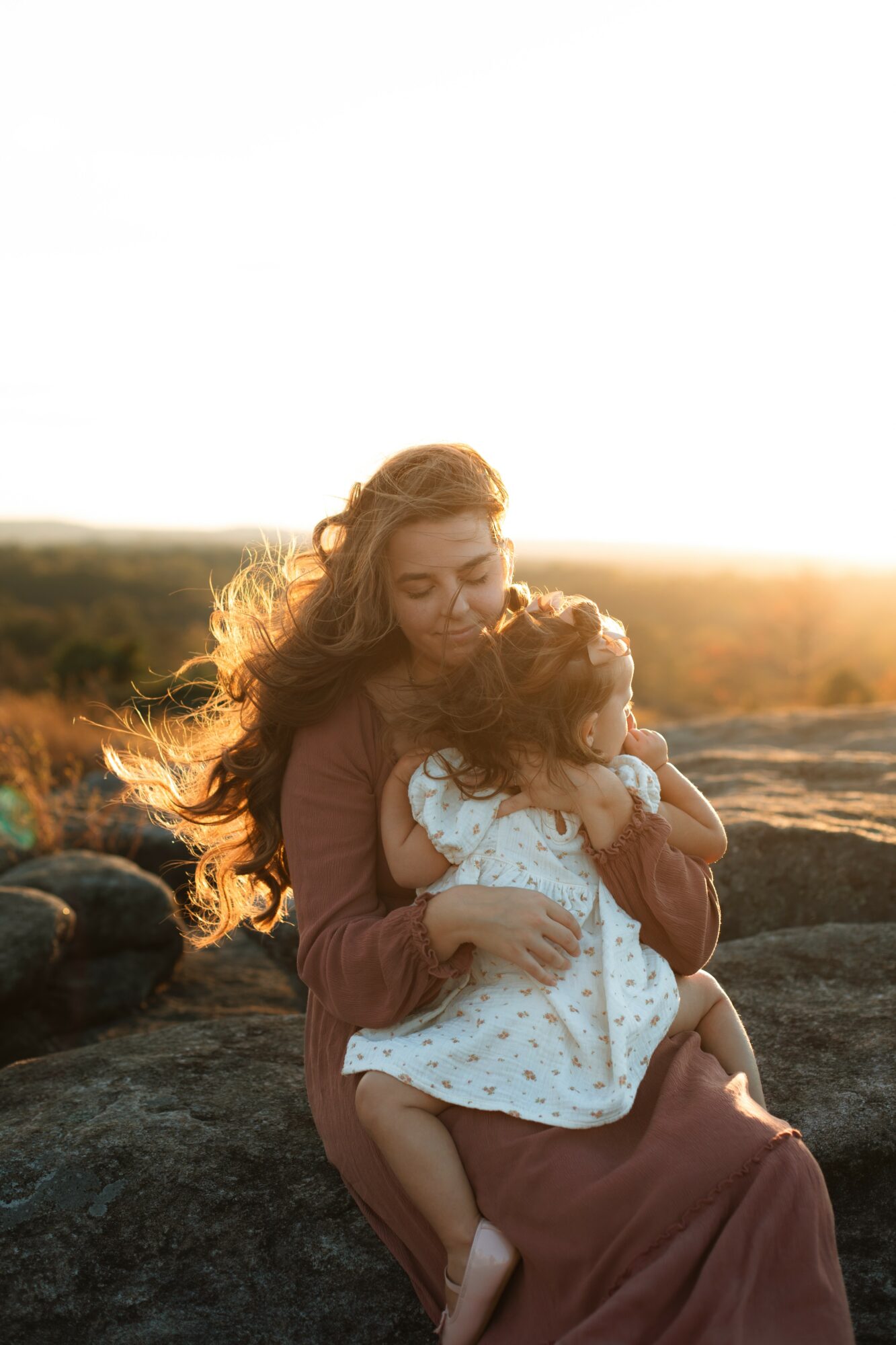 Woman holding a child outdoors during sunset, with a landscape background, warm lighting, and long hair.