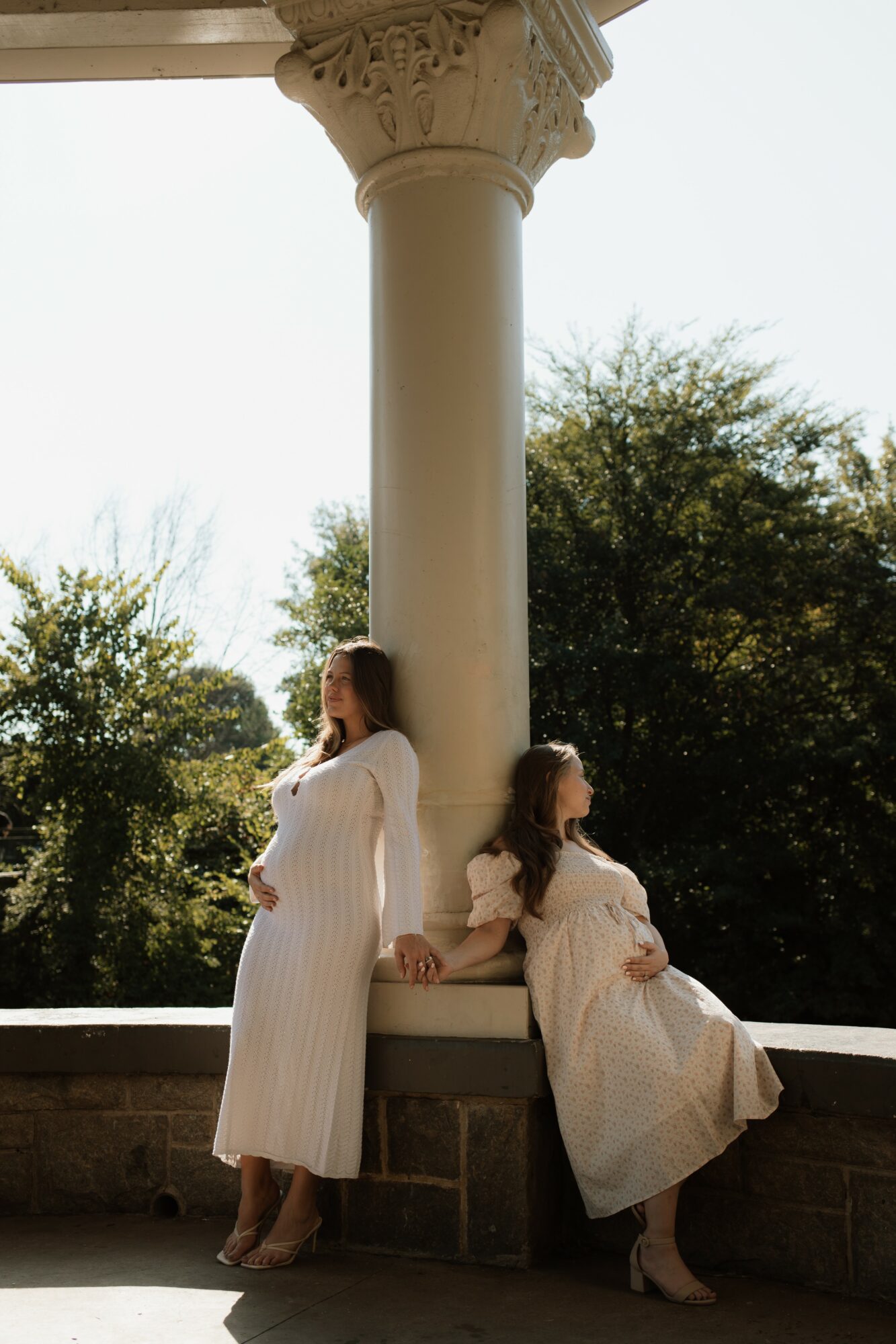 Two women in white dresses sit on a stone bench beside a large column, outdoors with trees in the background.