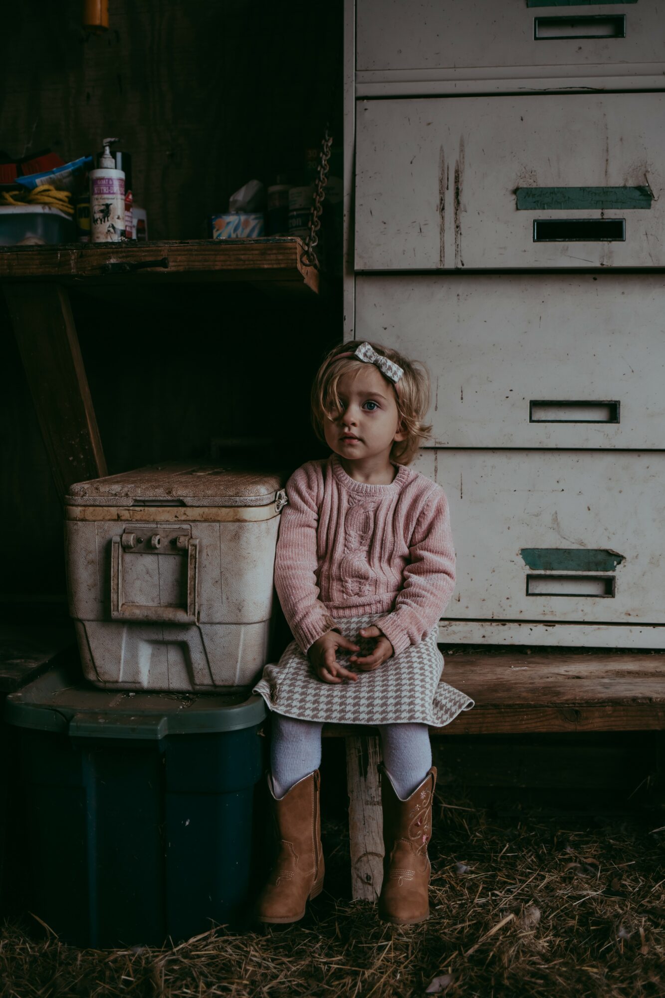 Young girl sitting on a small stool in front of a large white cabinet, wearing a pink sweater and brown boots.