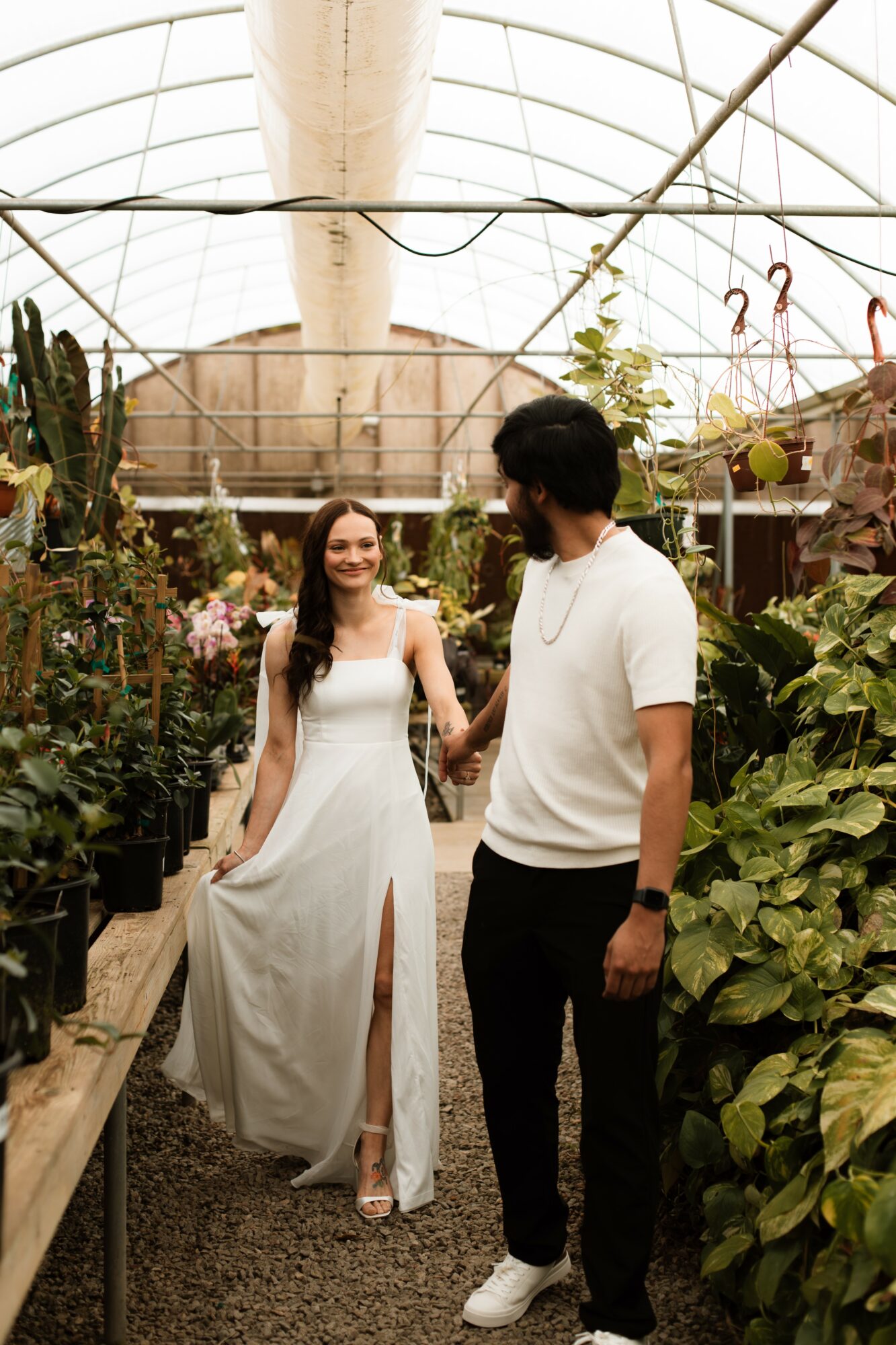 A woman in a white dress and a man in a white shirt and black pants stand in a greenhouse surrounded by plants, holding hands and smiling.
