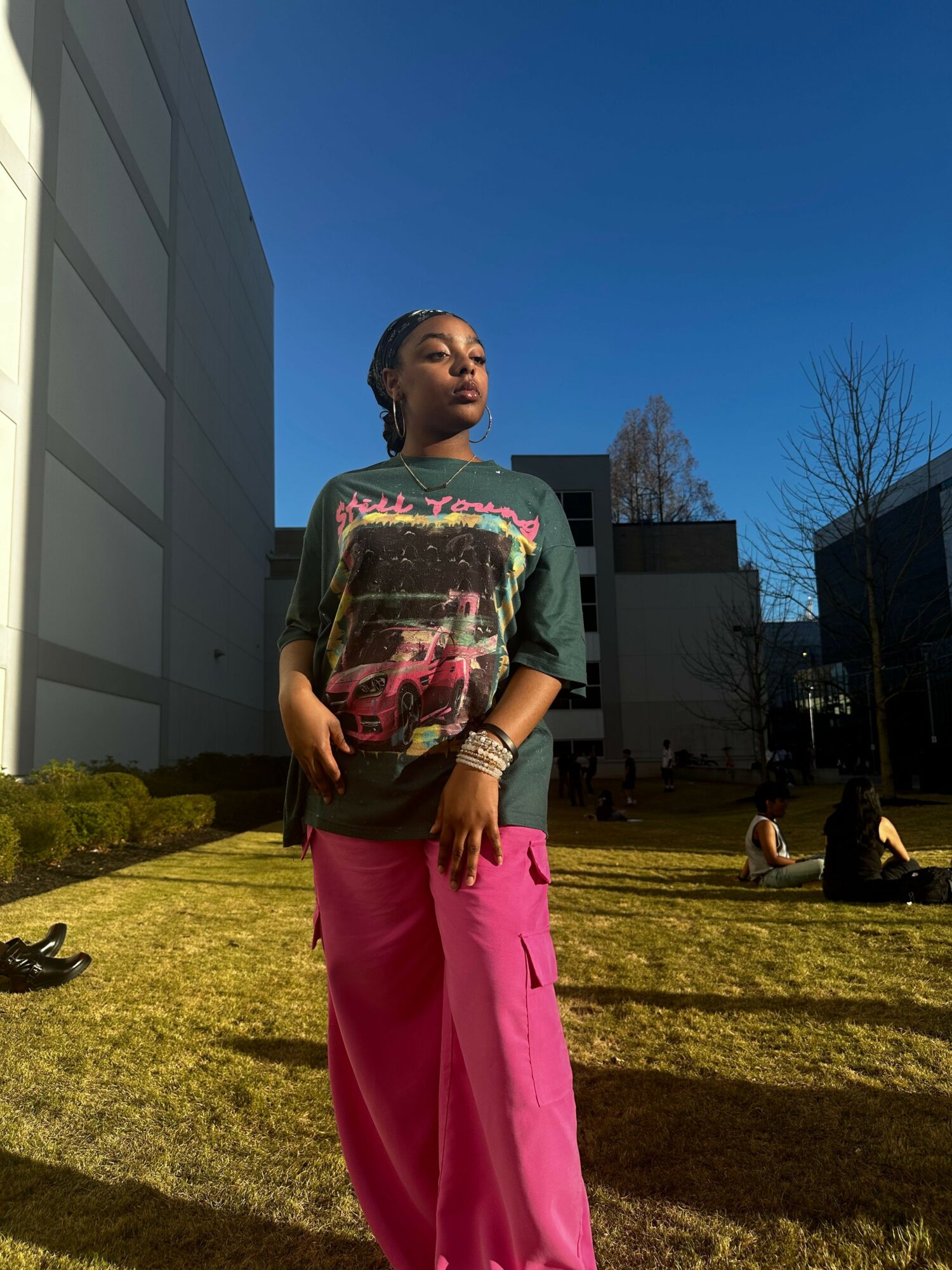 Person standing outdoors on grass, wearing a colorful top and pink pants, with a building and trees in the background.