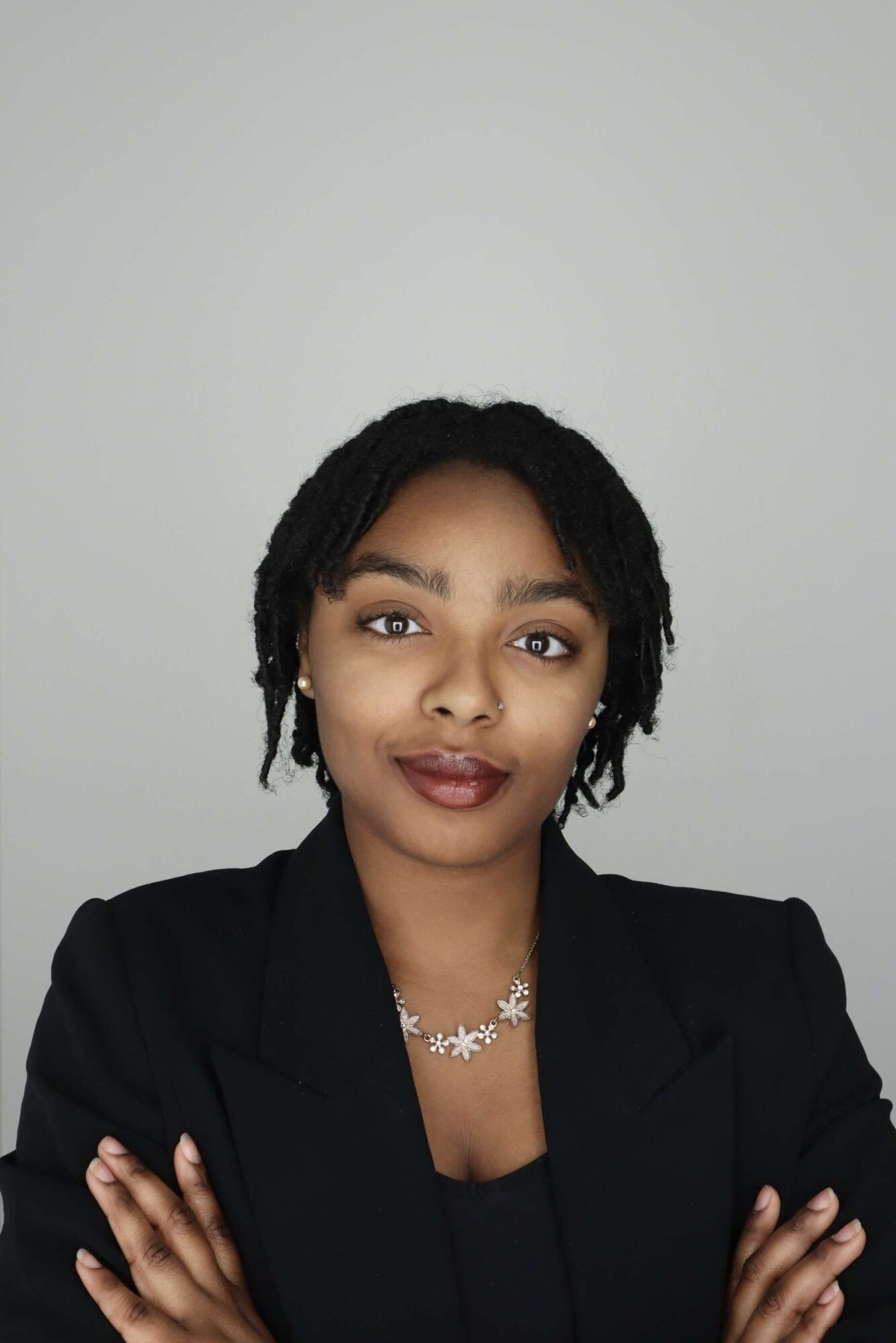 Portrait of a woman with short curly hair, wearing a black blazer and necklace, crossing arms, against a plain background.