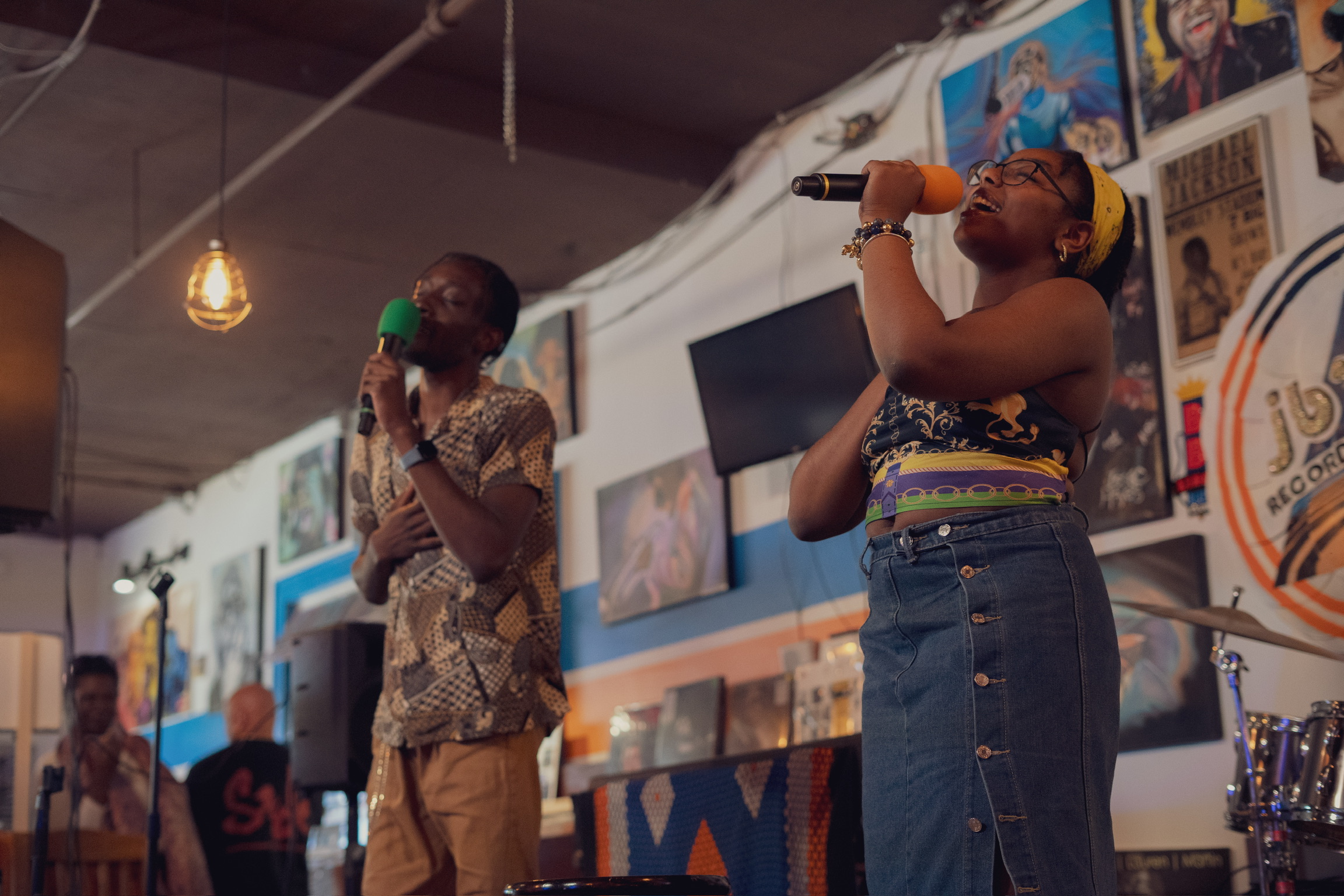 Two women singing into microphones on stage, one wearing a patterned dress and the other in a denim skirt and colorful top, in a decorated indoor venue.