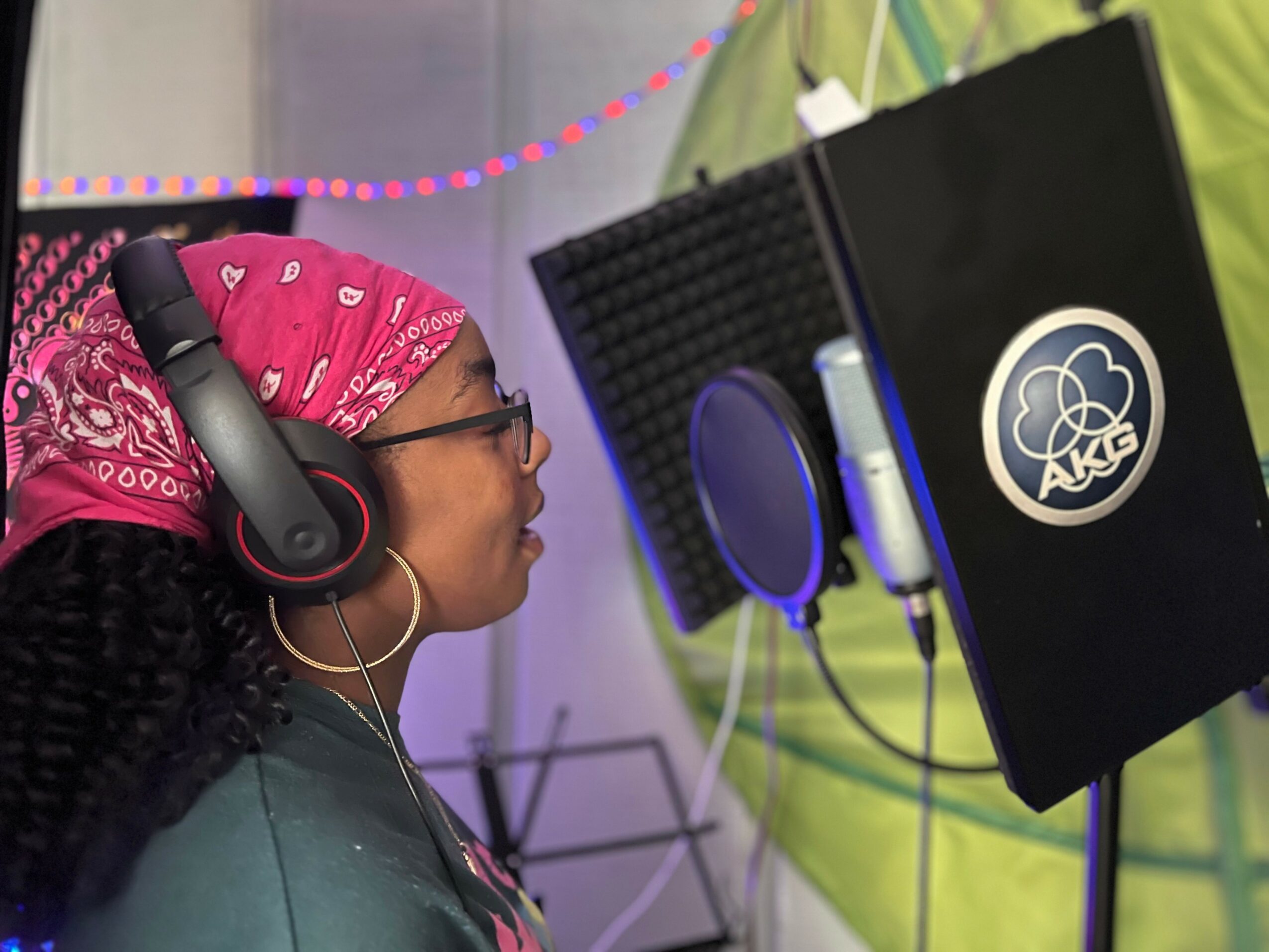 Woman wearing headphones and a pink bandana, recording vocals in a studio with a microphone and soundproofing.