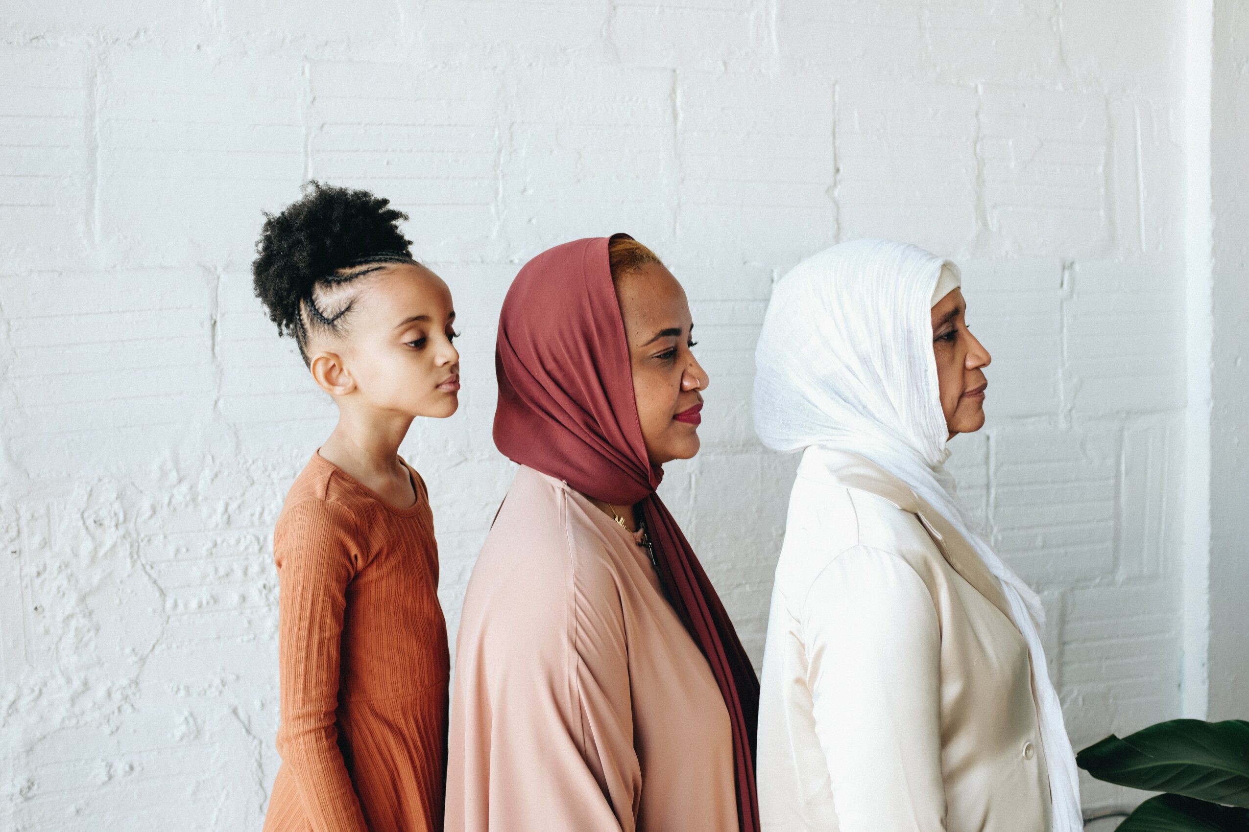 Three women standing side by side against a white brick wall, wearing head coverings and modest clothing.