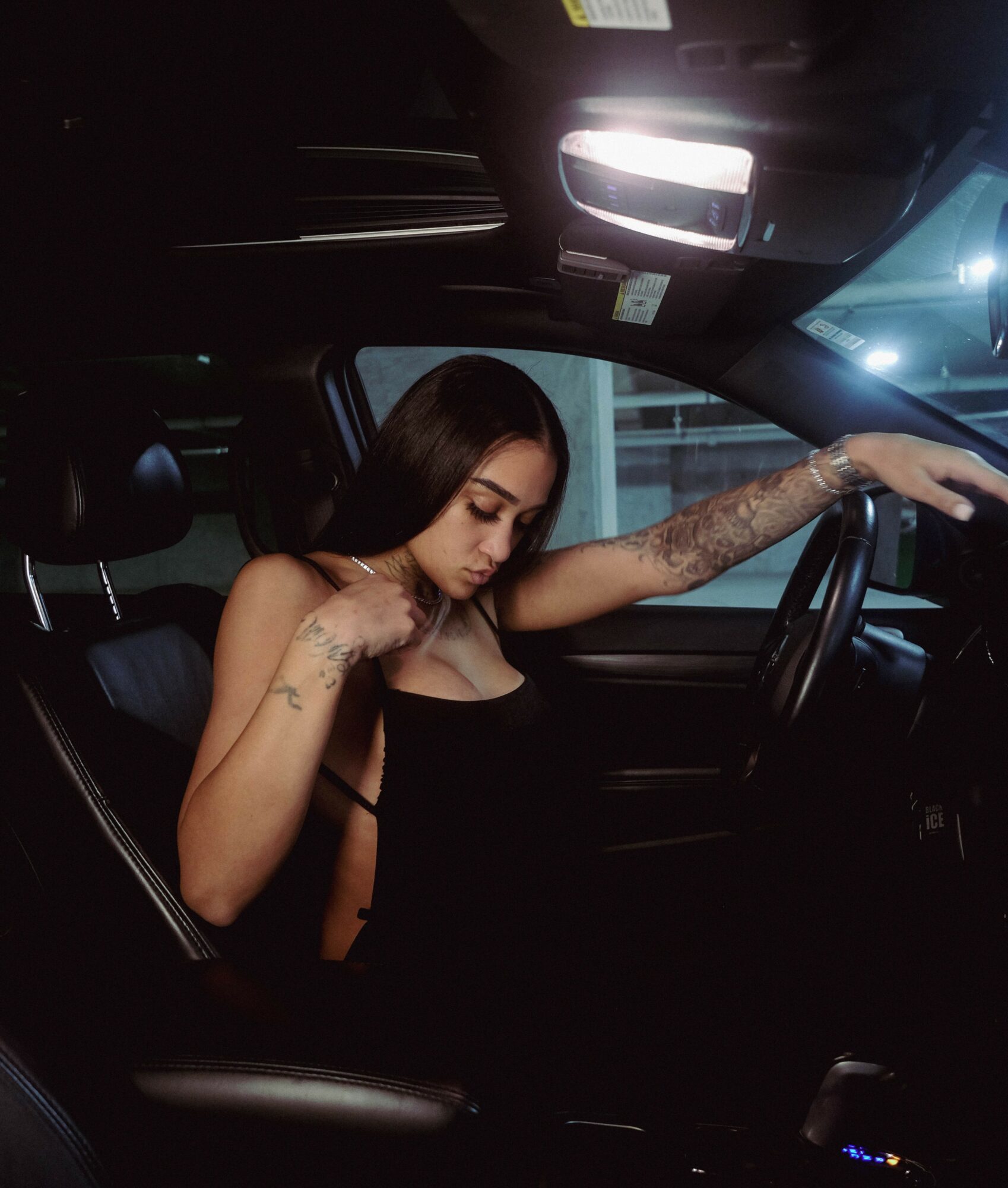 Young woman with long dark hair sitting in a car, looking down, wearing a black top, with tattoos on her arm.