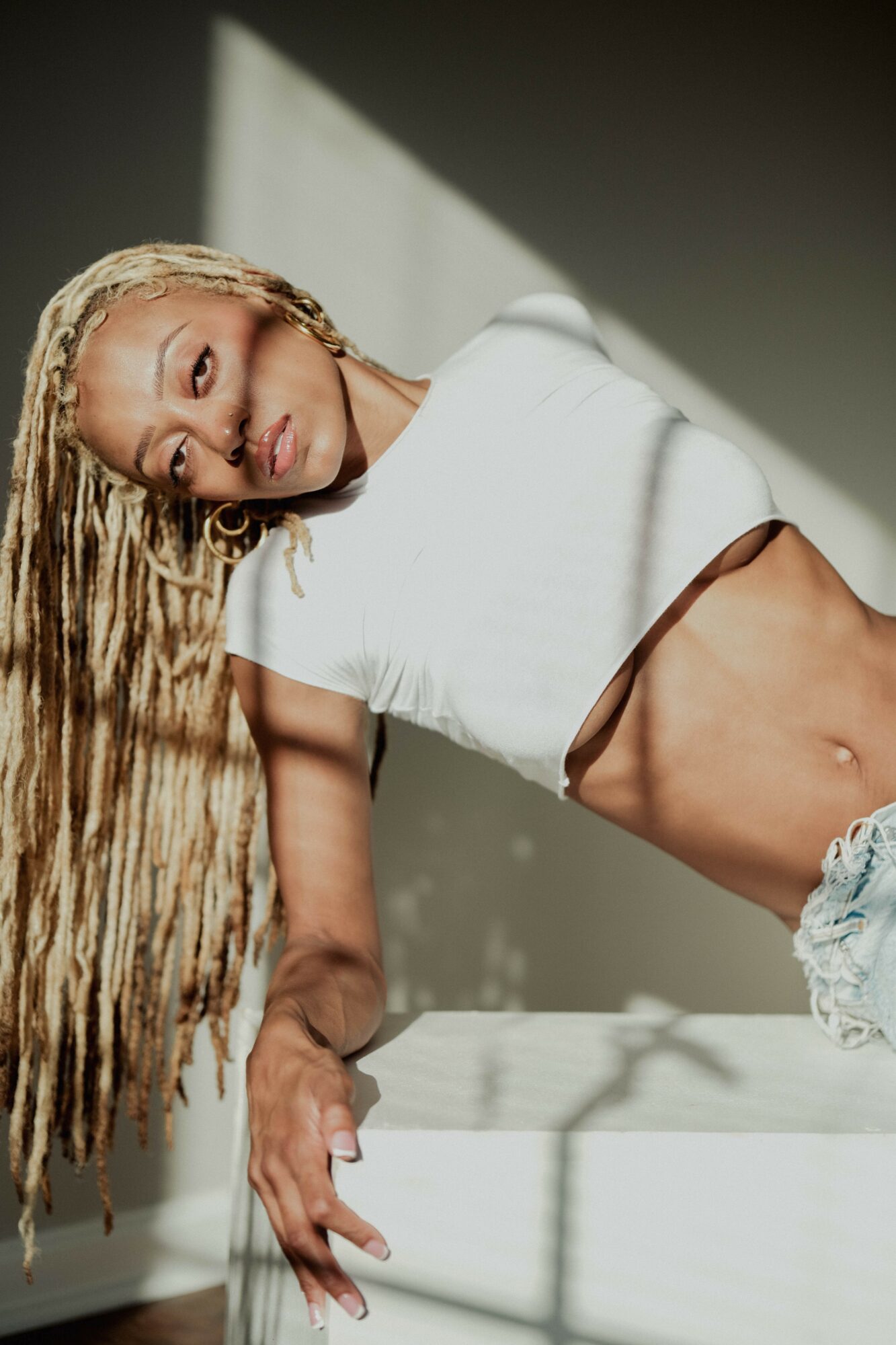 Young woman with long braided hair lying on her side, wearing a white crop top and denim shorts, with sunlight and shadows.