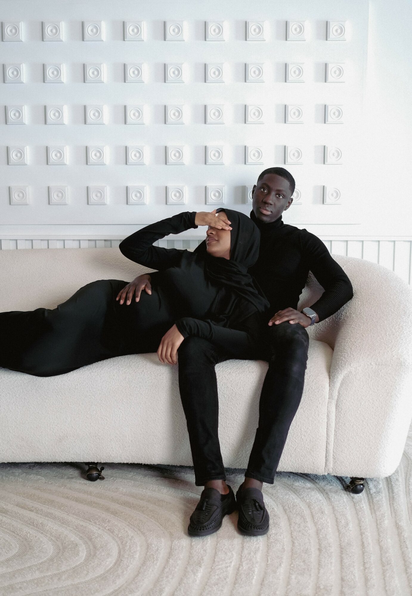 Person sitting on a white sofa, wearing black clothing, with a wall of white objects behind them.