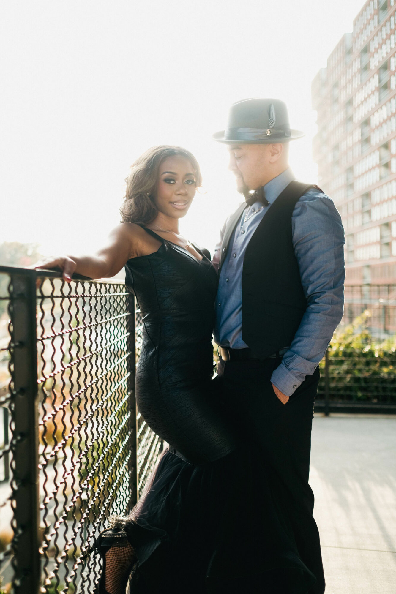 A woman and a man stand outdoors near a railing, with buildings in the background, sunlight brightening the scene.