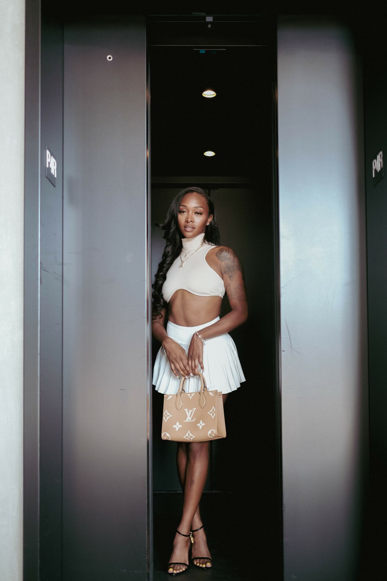 Woman standing in an elevator, wearing a white crop top and skirt, holding a beige bag, with dark hair and heels.