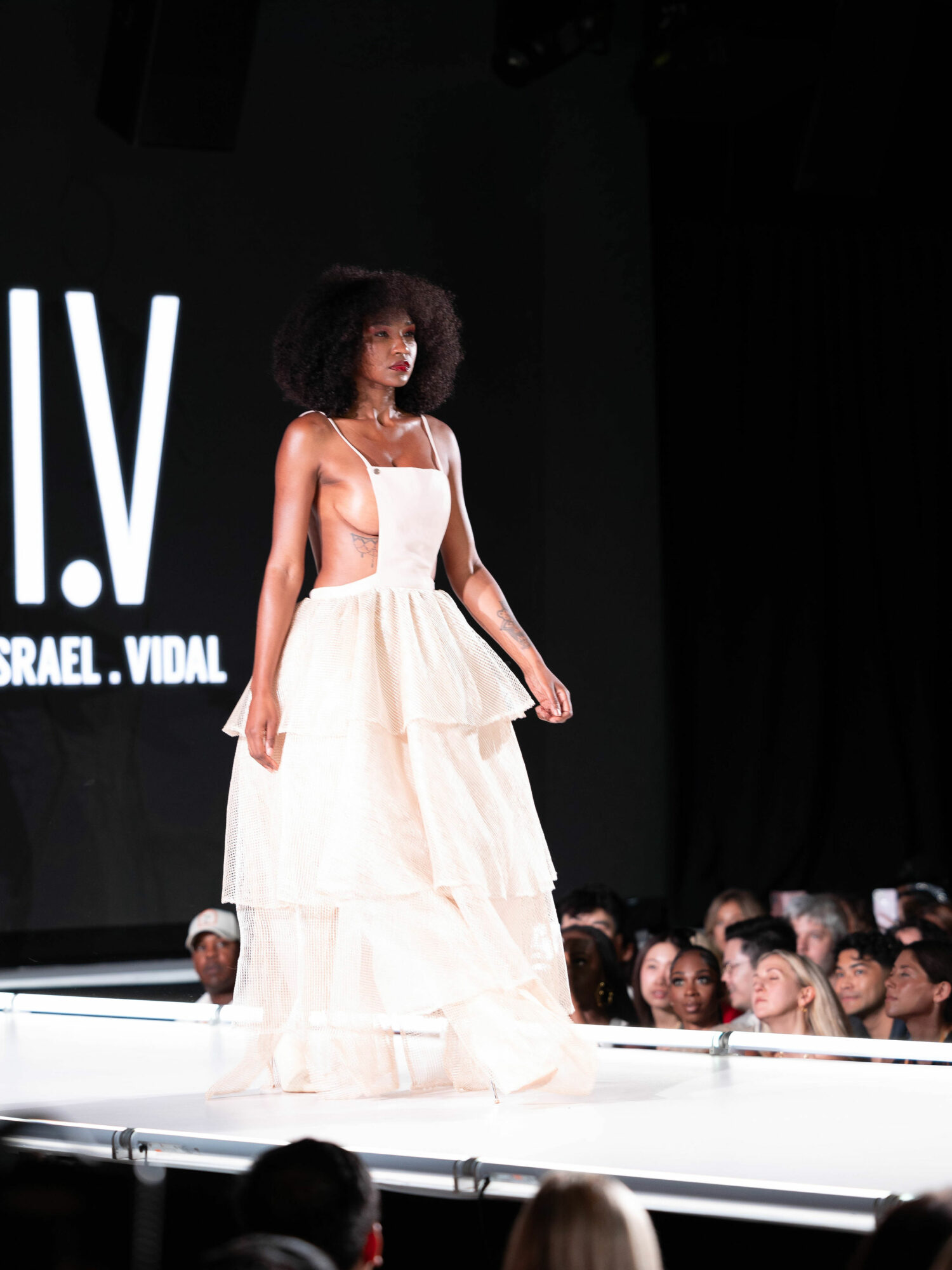 Model walking on runway in a white dress with audience in background, stage with black backdrop, and text on screen.