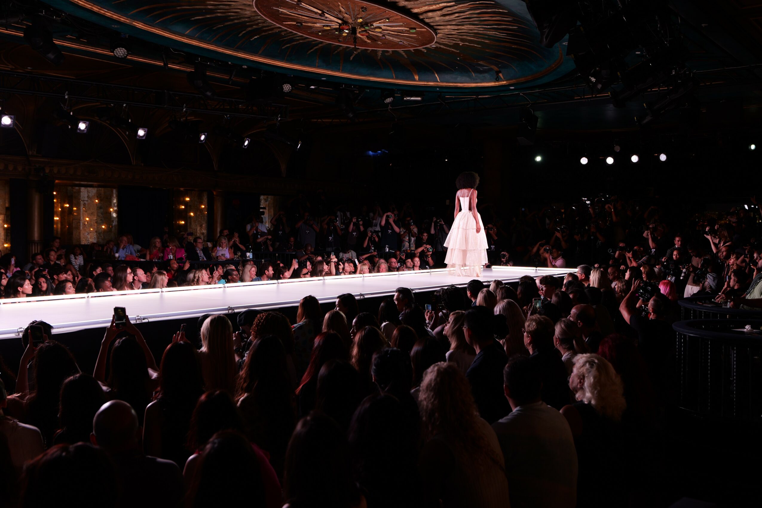 A model walks down a runway with an audience on both sides, in a dimly lit venue with a decorated ceiling.