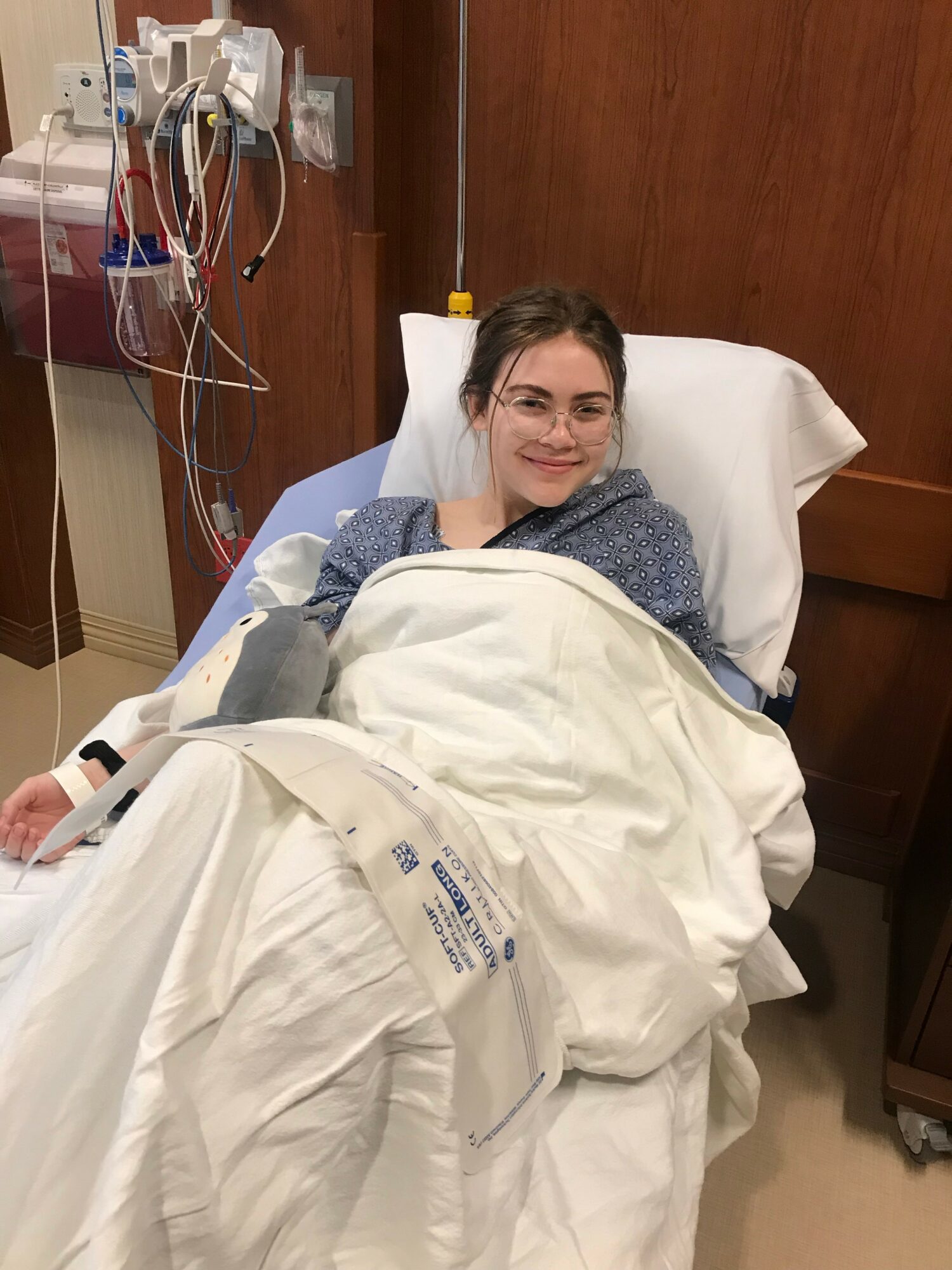 Young woman in hospital bed smiling, covered with white blanket, medical equipment nearby, wooden wall behind her.