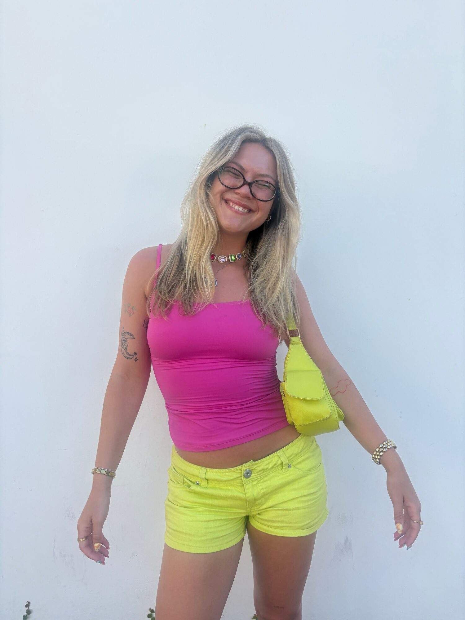 Woman with long blonde hair wearing a pink top and yellow shorts, smiling, standing against a plain background.