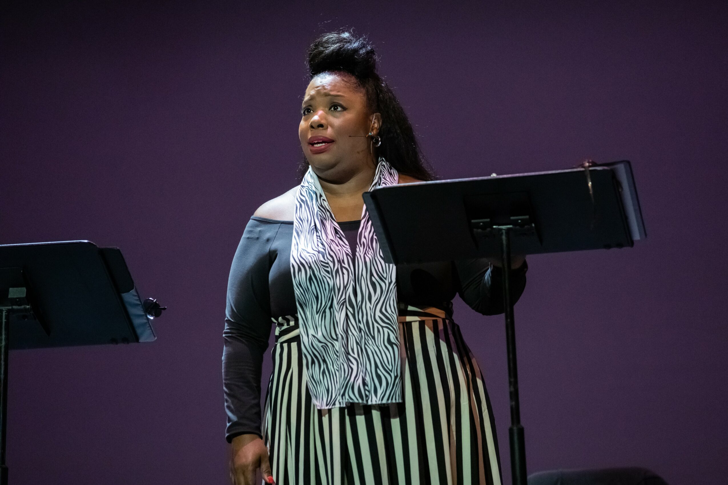 Woman standing behind a music stand on stage with a purple background, wearing a striped skirt and patterned scarf.