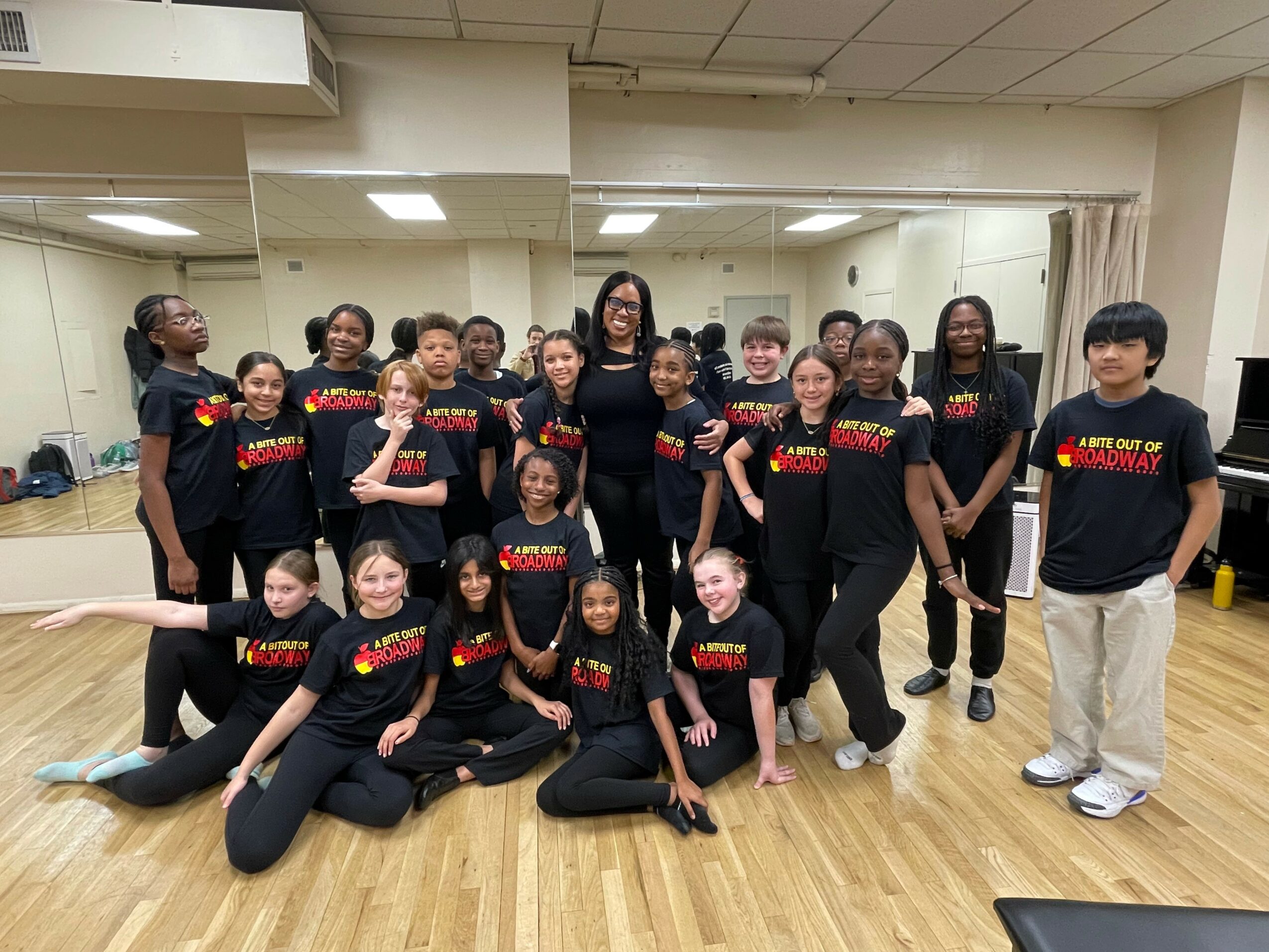 Group of young dancers in black outfits with red and yellow text, posing in a dance studio with mirrors and wooden floor.