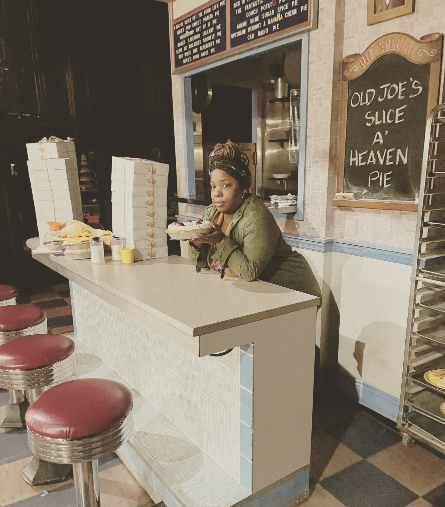 Woman leaning on a counter inside a bakery or cafe, with a chalkboard sign reading 'Old Joe's Slice A Heaven Pie' behind her.