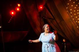 A woman singing passionately on stage with red lights and a wooden backdrop.