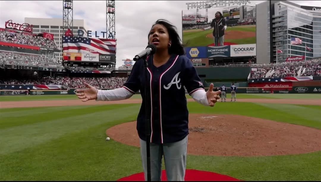 Person standing on baseball field with arms outstretched, stadium and scoreboard in background.