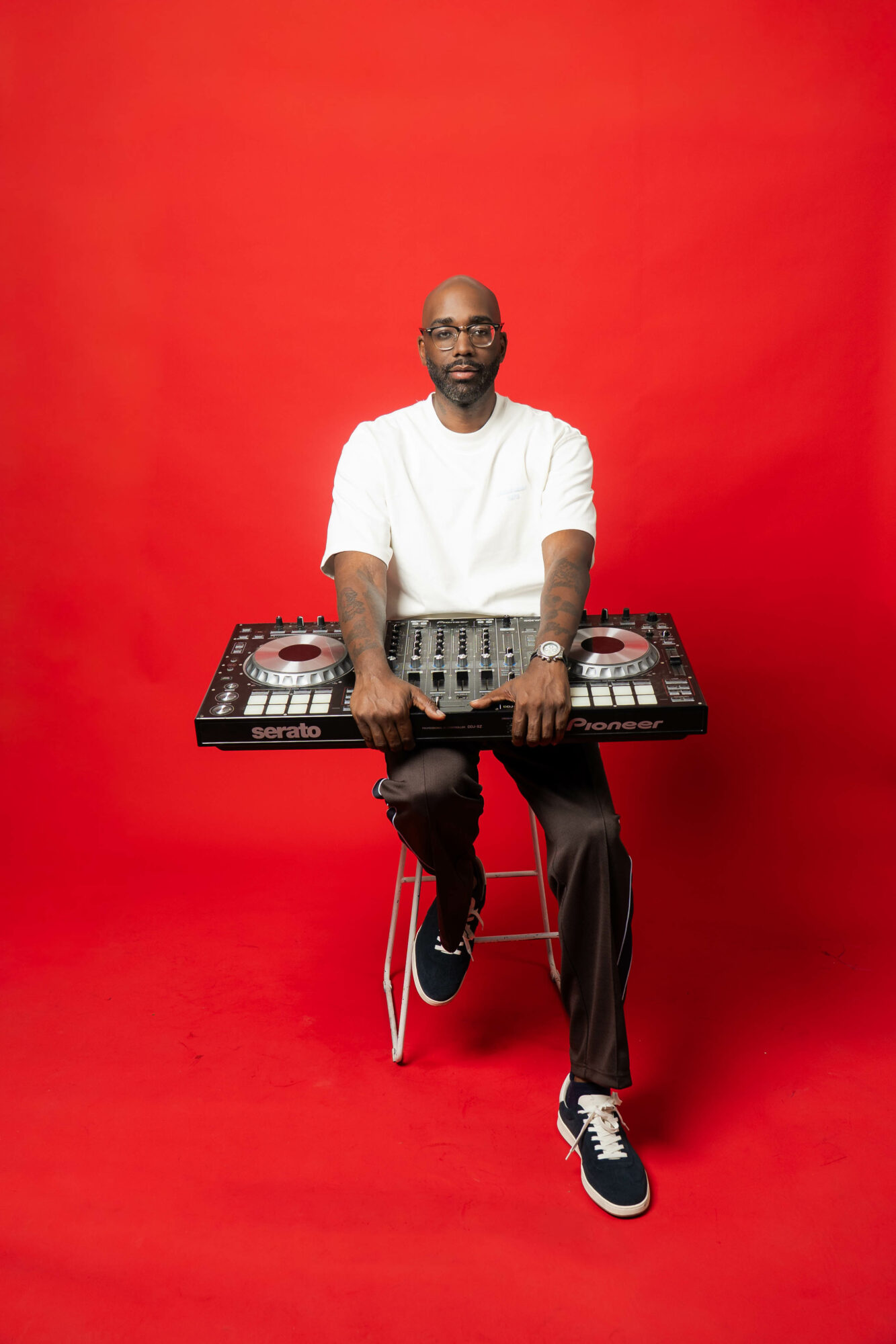 Man standing behind DJ equipment against a red background, wearing a white shirt and black pants.