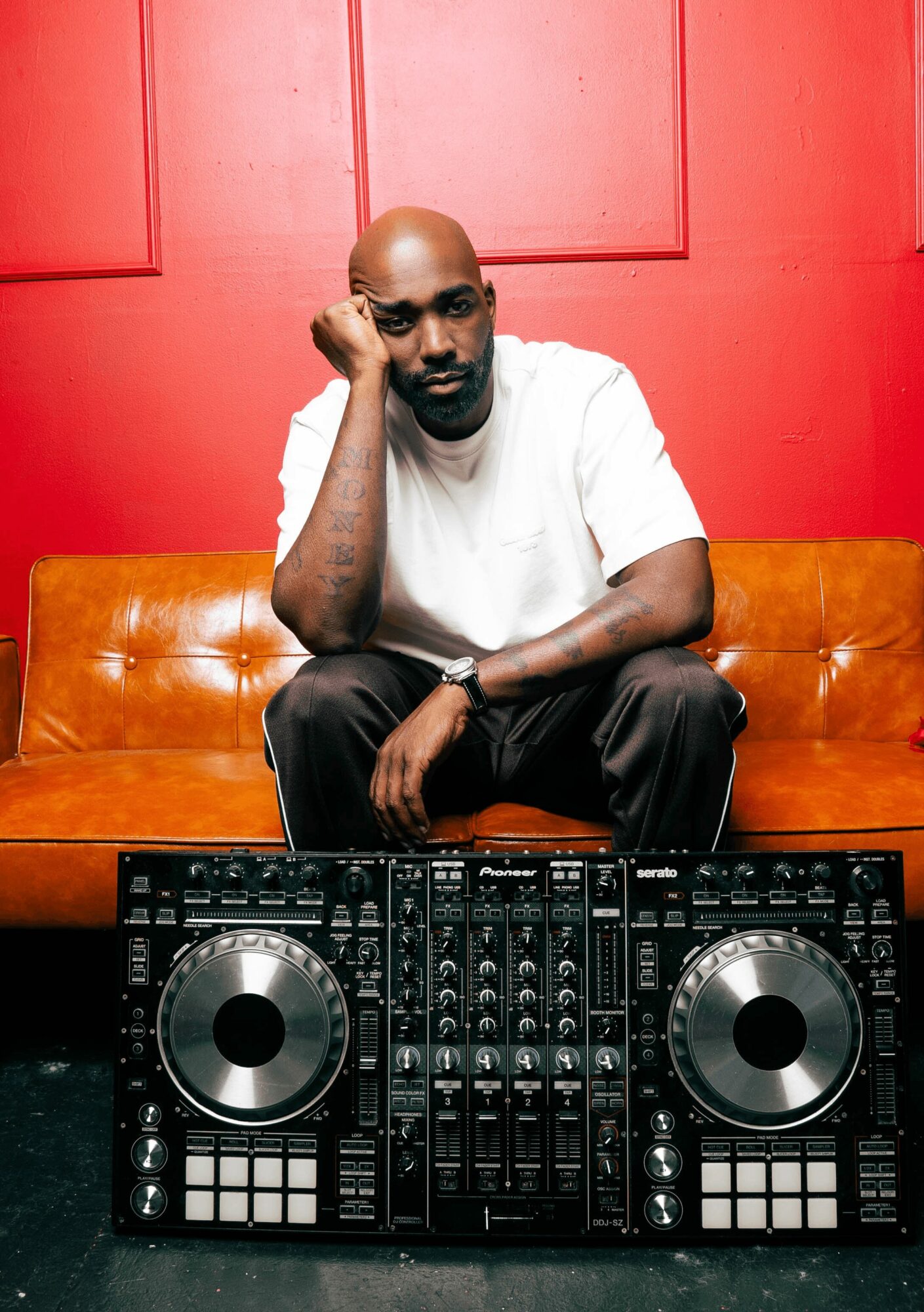 Man sitting on a sofa with DJ equipment in front, red wall background, resting his head on his hand.
