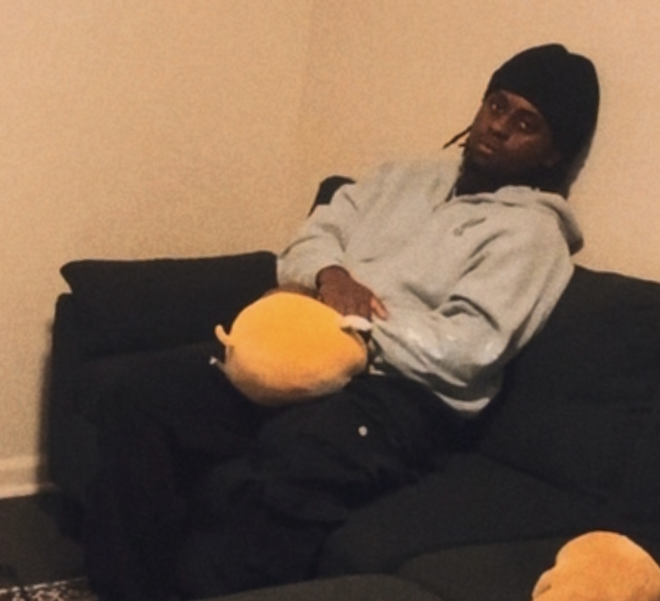 Person sitting on a black couch holding a plush toy, wearing a gray hoodie and black beanie, in a room with beige walls.