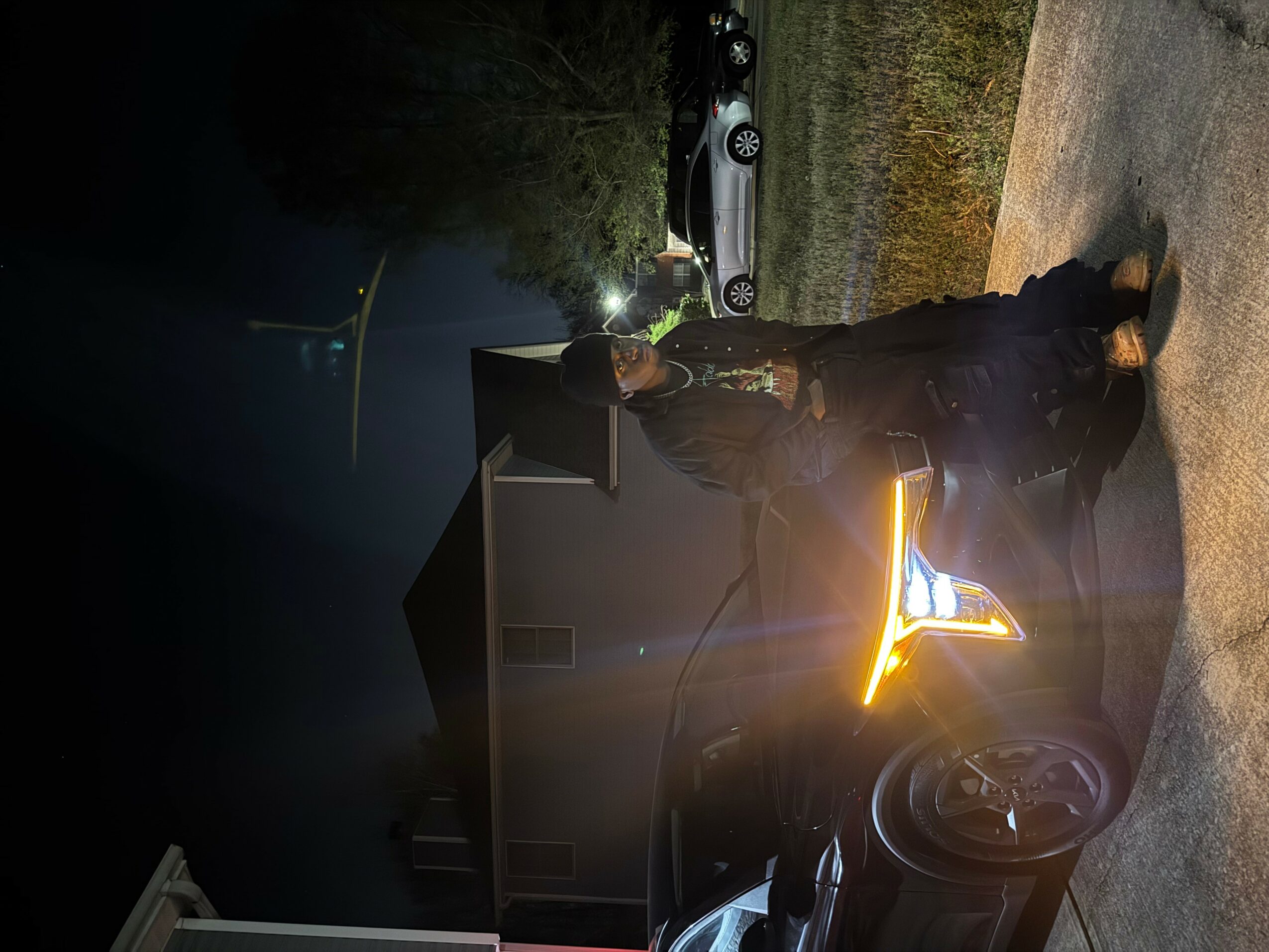 Person standing outside near a house at night, with cars parked in the background, illuminated by a bright light.
