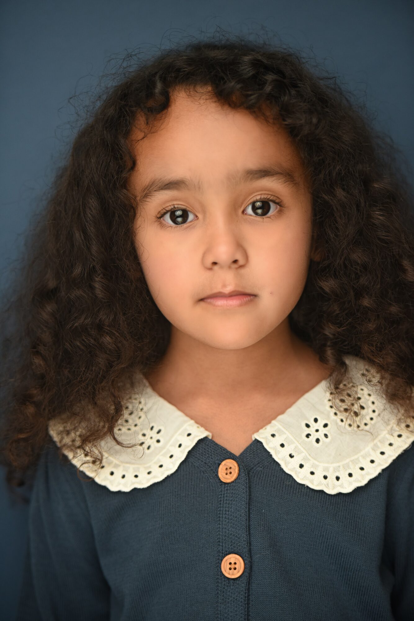Young girl with curly hair wearing a navy blue top with a white lace collar, looking directly at the camera.