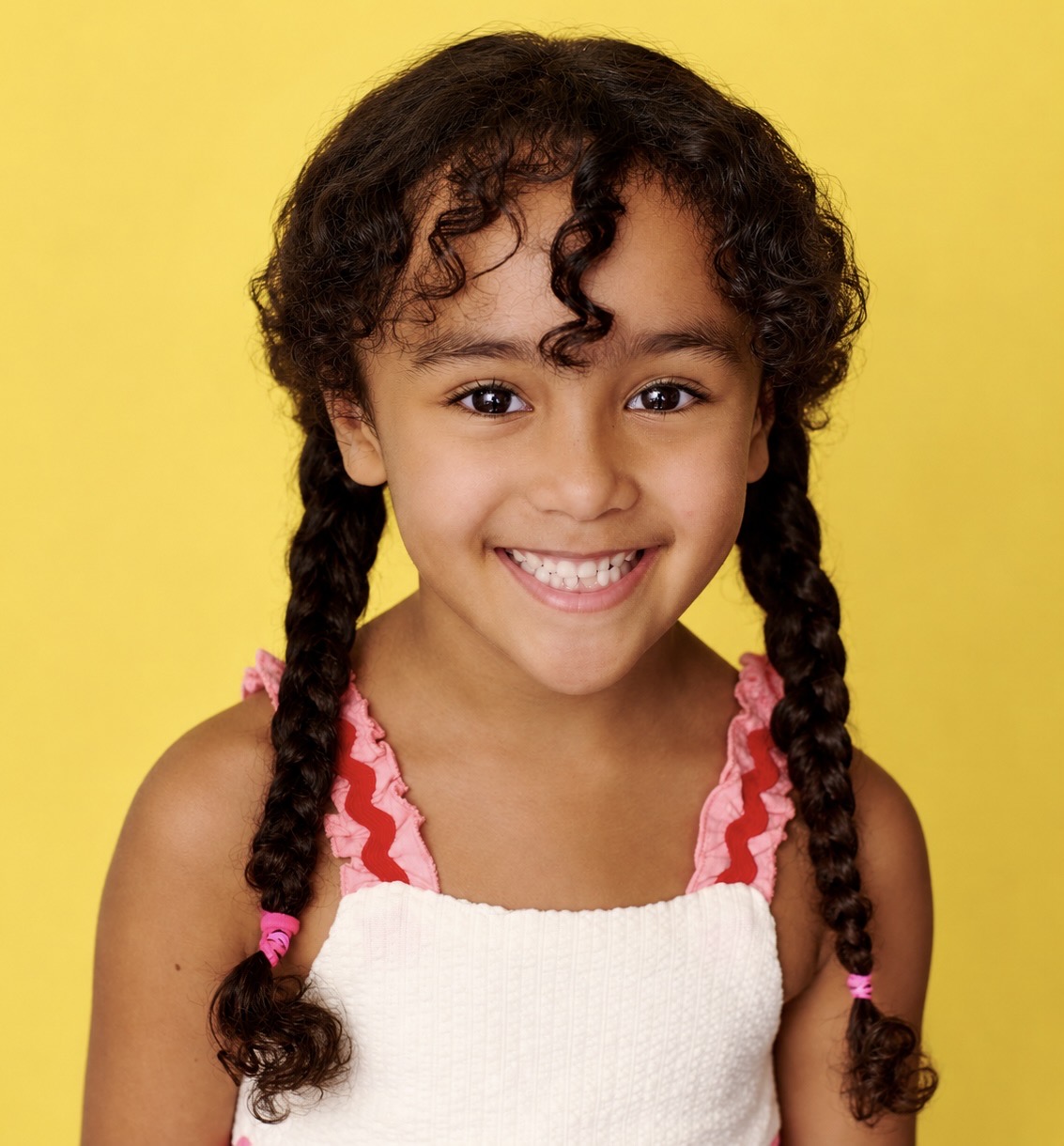 Smiling girl with curly hair in braids, wearing a white top with pink ruffles, against a yellow background.