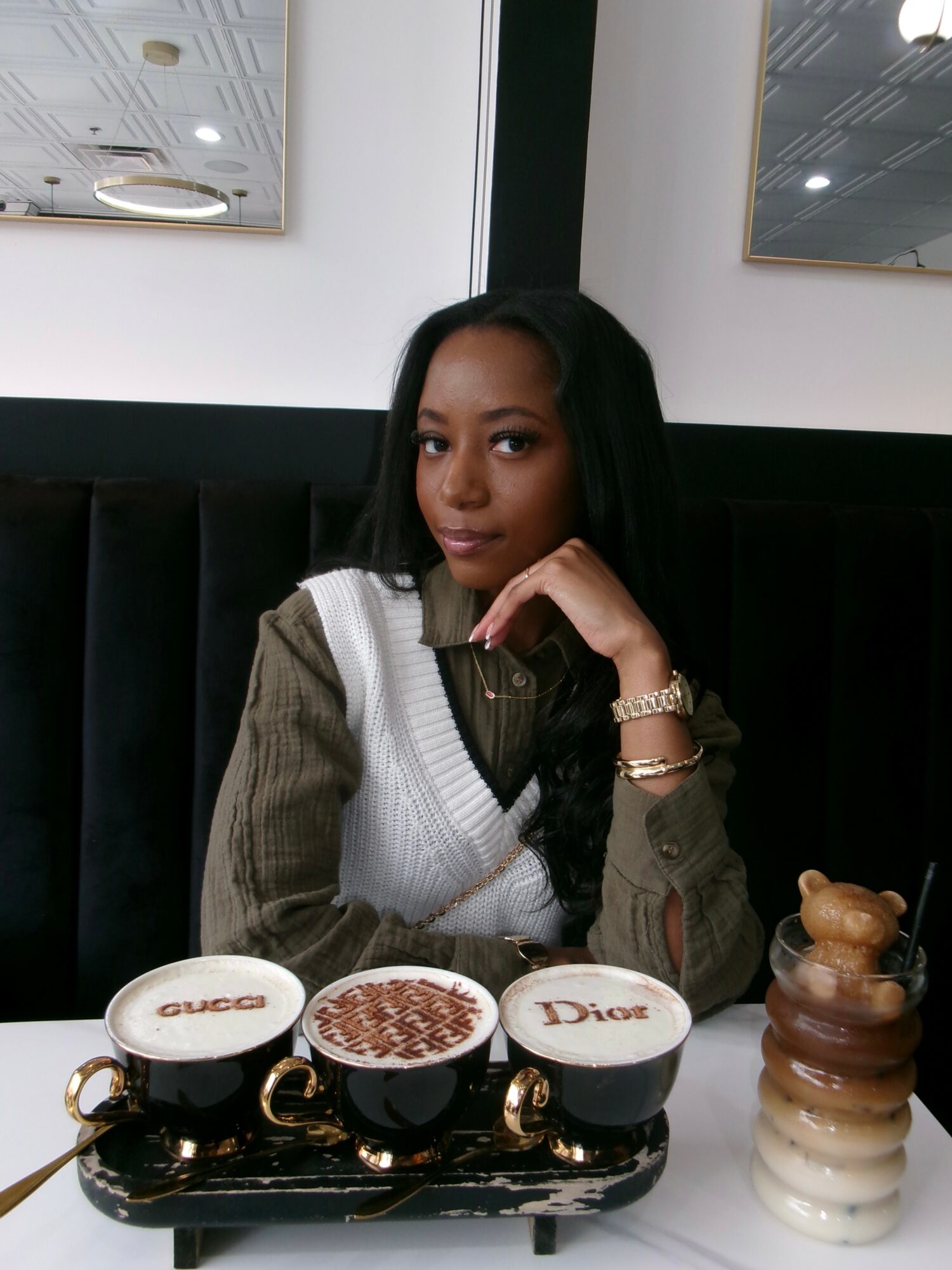 Woman sitting at a table with three small Gucci, Dior, and chocolate-themed containers, and a stack of layered desserts, in a modern setting.