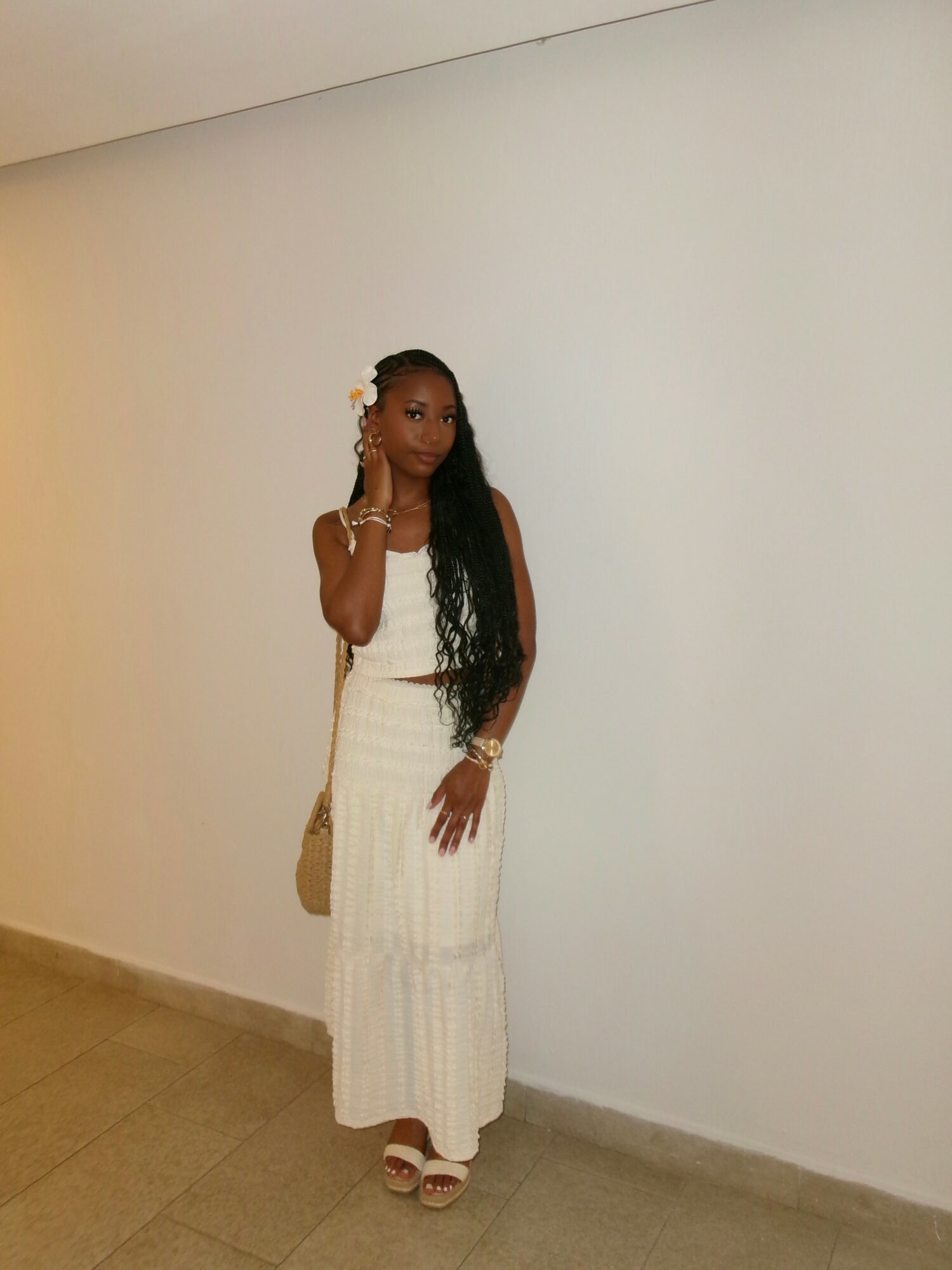 Woman in white dress with long dark hair standing against a plain white wall, holding a small bag, wearing sandals.