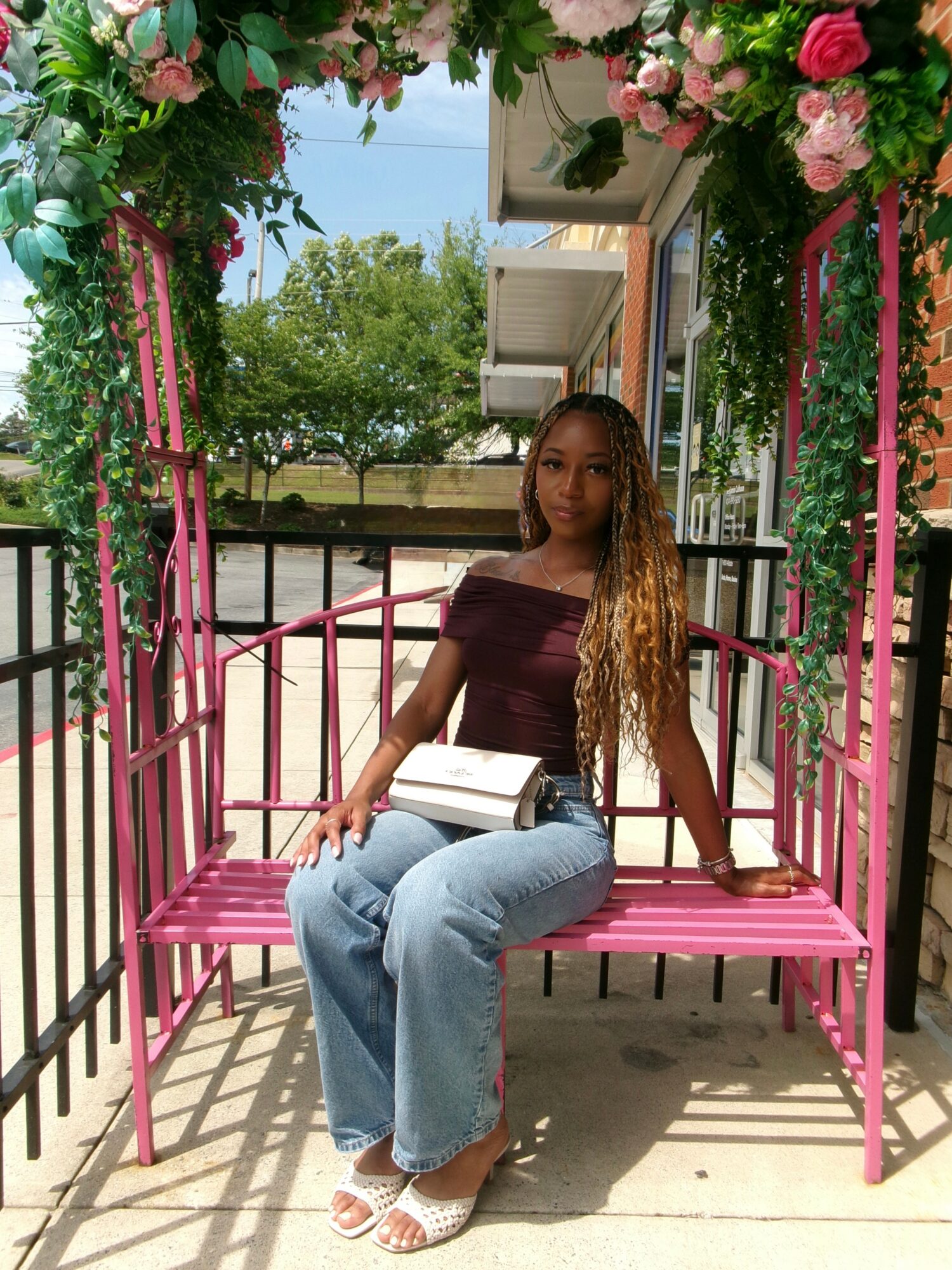 Young woman sitting on a pink swing with floral arch, outdoor setting, trees, and building in background.