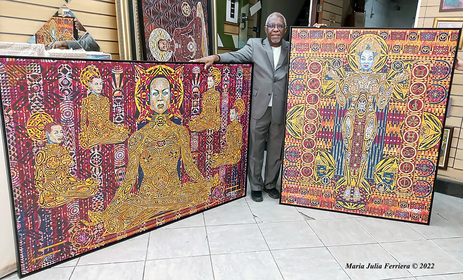 Man standing between two colorful, intricate paintings with multiple figures, in an art gallery setting.
