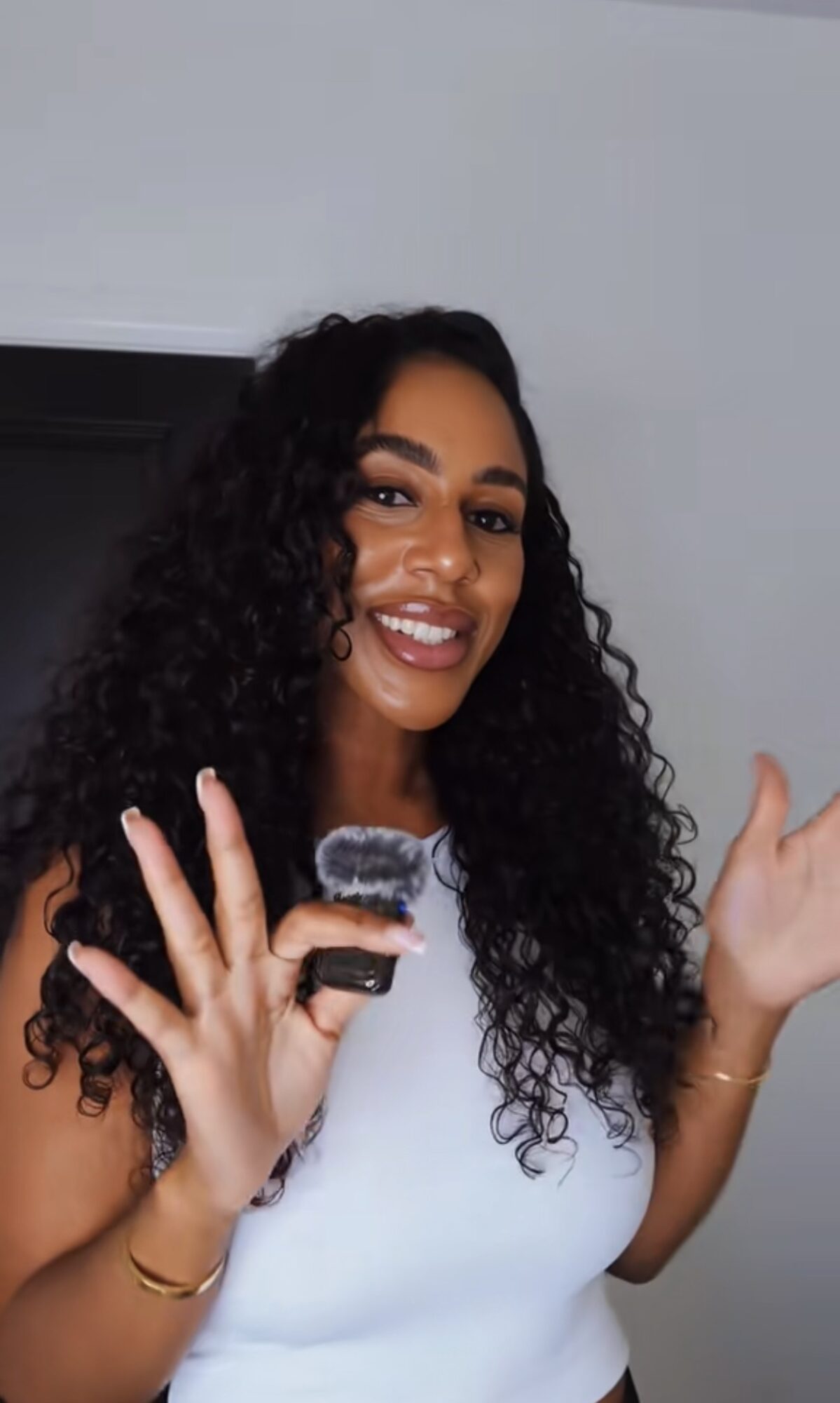 Smiling woman with curly hair holding a microphone, wearing a white top, against a plain background.