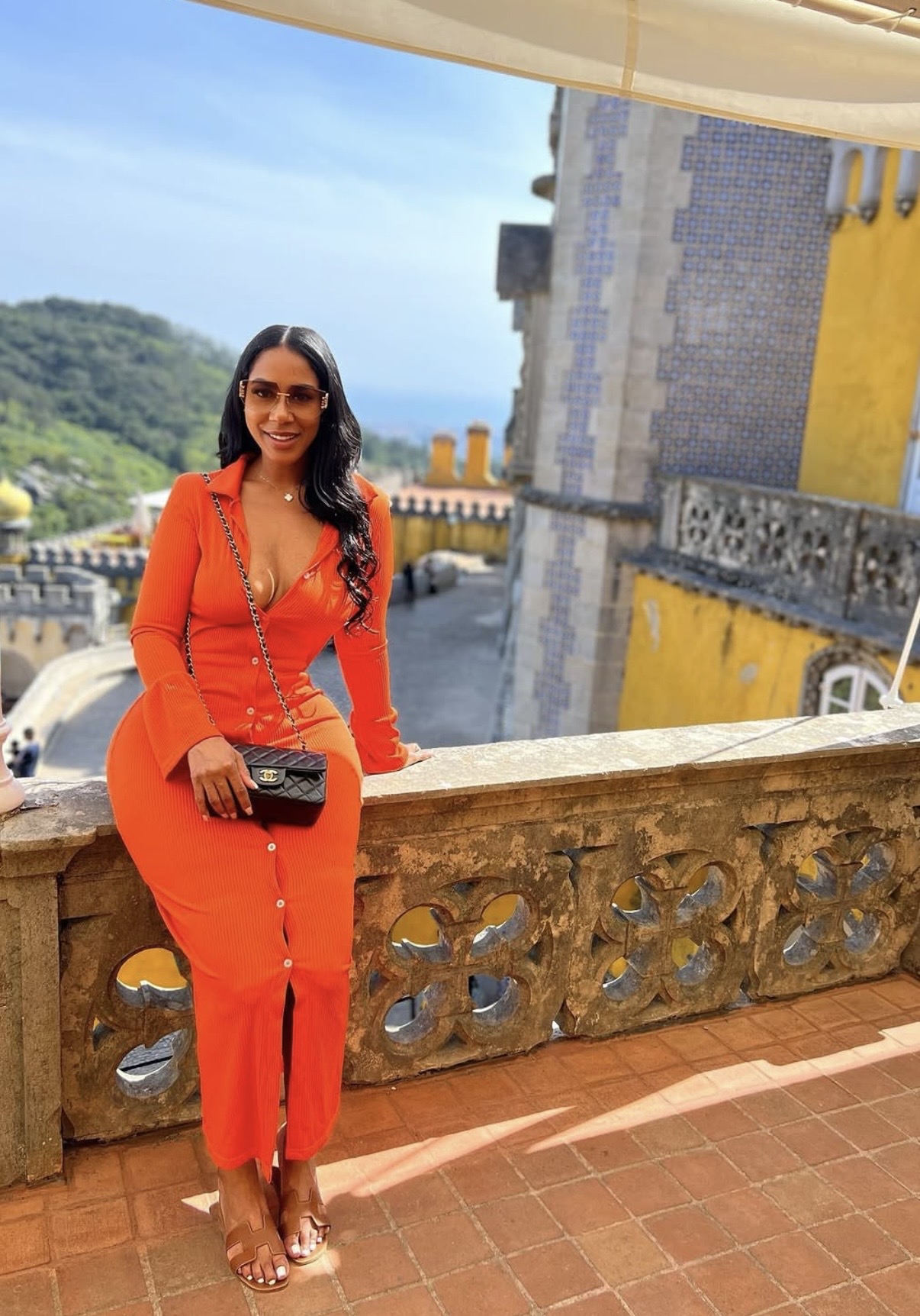 Woman in orange dress and sunglasses standing on a balcony with a scenic background, holding a black purse.