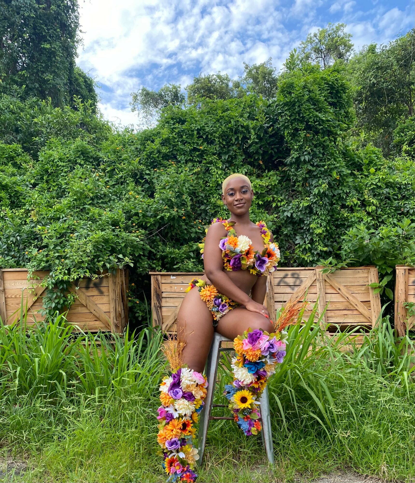 Person sitting outdoors on a chair, surrounded by greenery and wearing a colorful floral outfit, smiling at the camera.