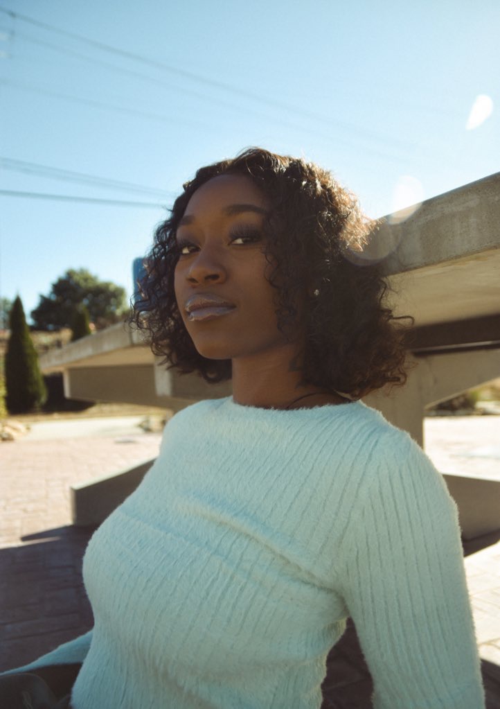 Young woman with curly hair wearing a light-colored sweater outdoors, leaning against a wooden structure, sunny day.
