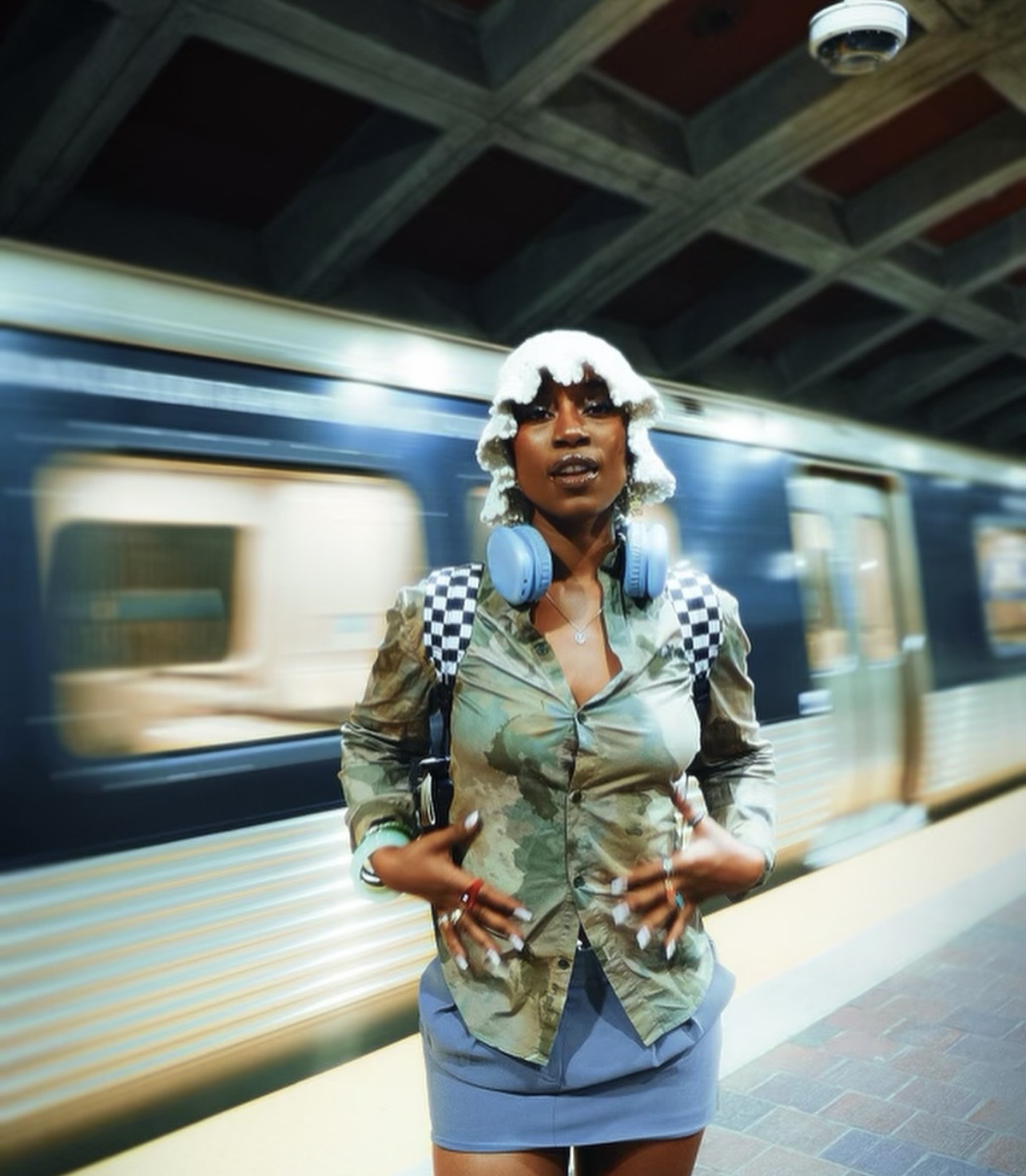 Woman with headphones and checkered backpack standing on subway platform, train passing by in background.