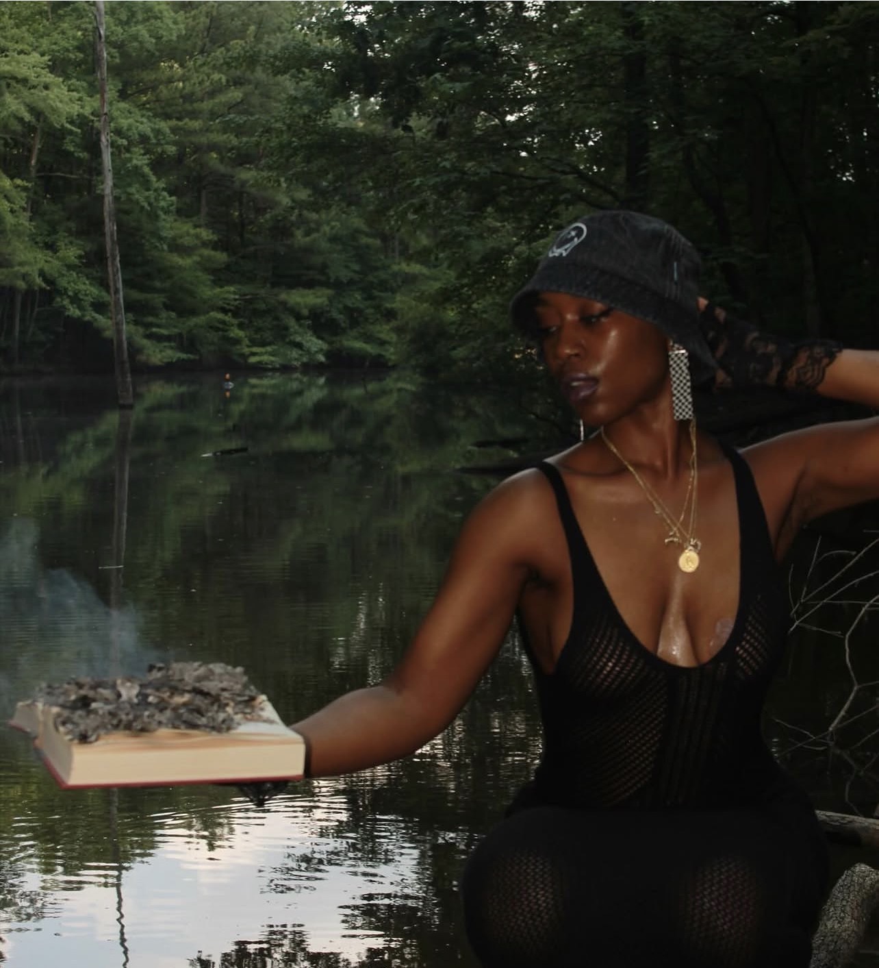 Woman in black outfit and hat sitting by a river with trees in the background, holding a book, looking to the side.