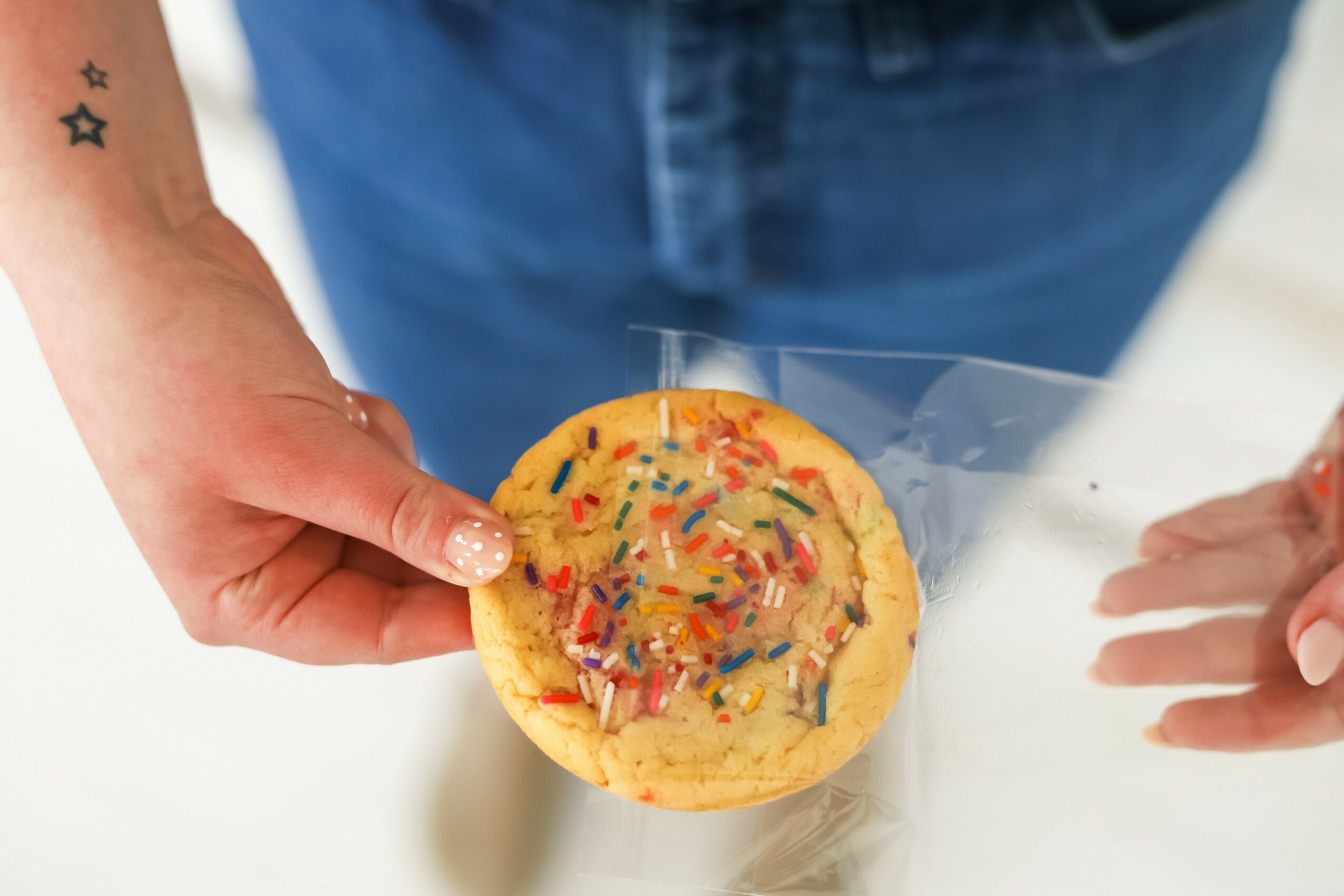 Person holding a decorated cookie with colorful sprinkles, with another hand reaching towards it, against a blurred background.