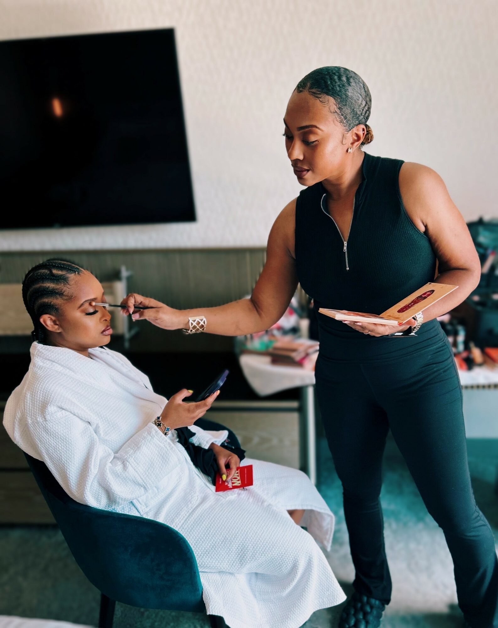 Two women in a room, one sitting in a chair with a phone, the other standing and handing her a makeup palette.