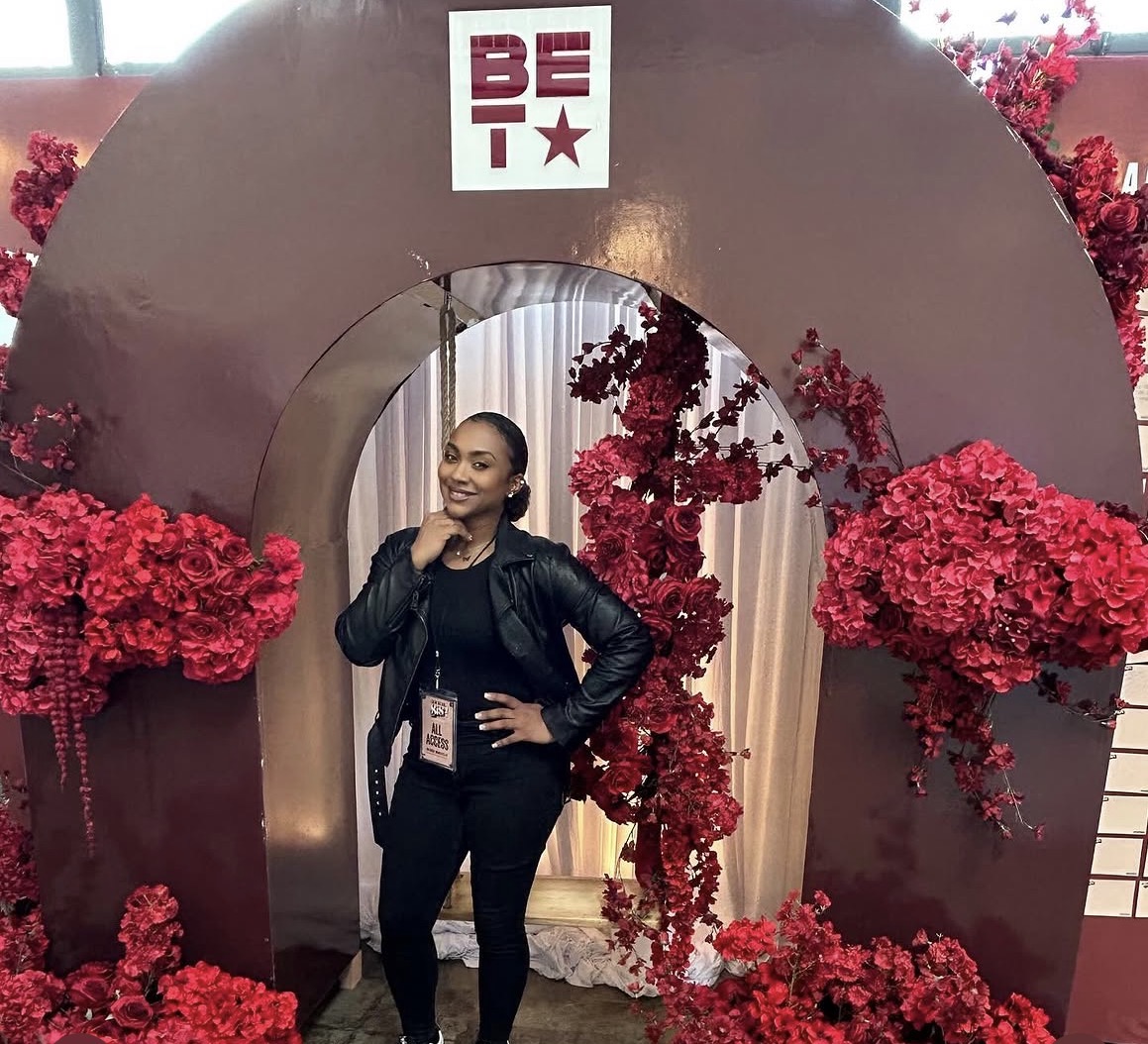 Woman standing in front of a decorative arch with red flowers and a BET logo, smiling with hand on hip.