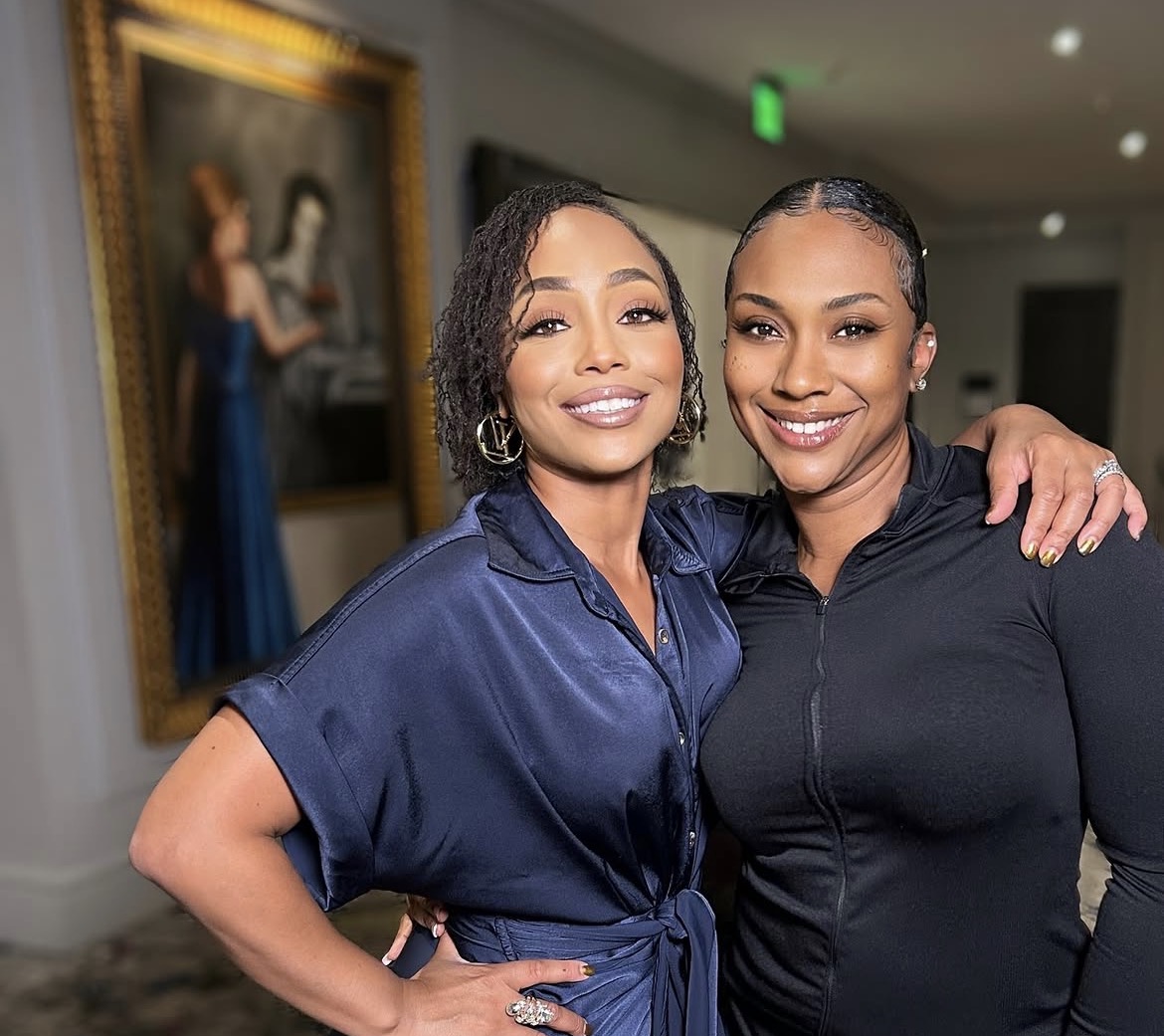 Two women smiling and posing together indoors, one with short curly hair and the other with straight hair, in front of a painting.
