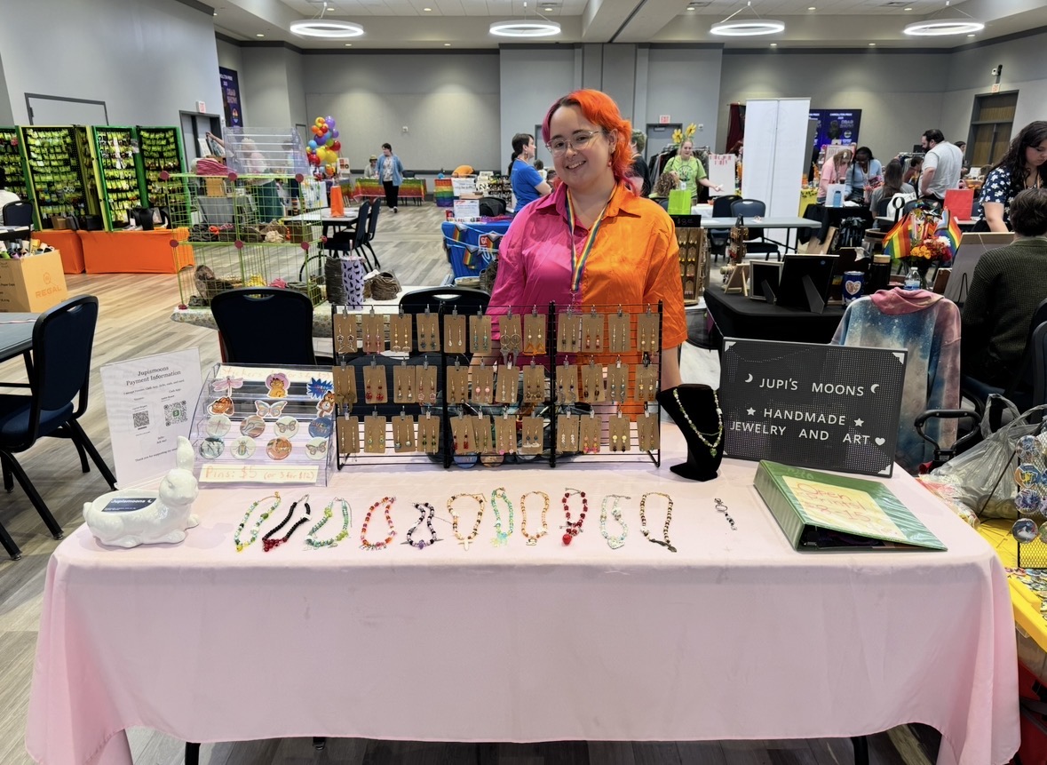 Woman with orange hair and pink and orange clothing stands behind a table with jewelry displays at an indoor event.