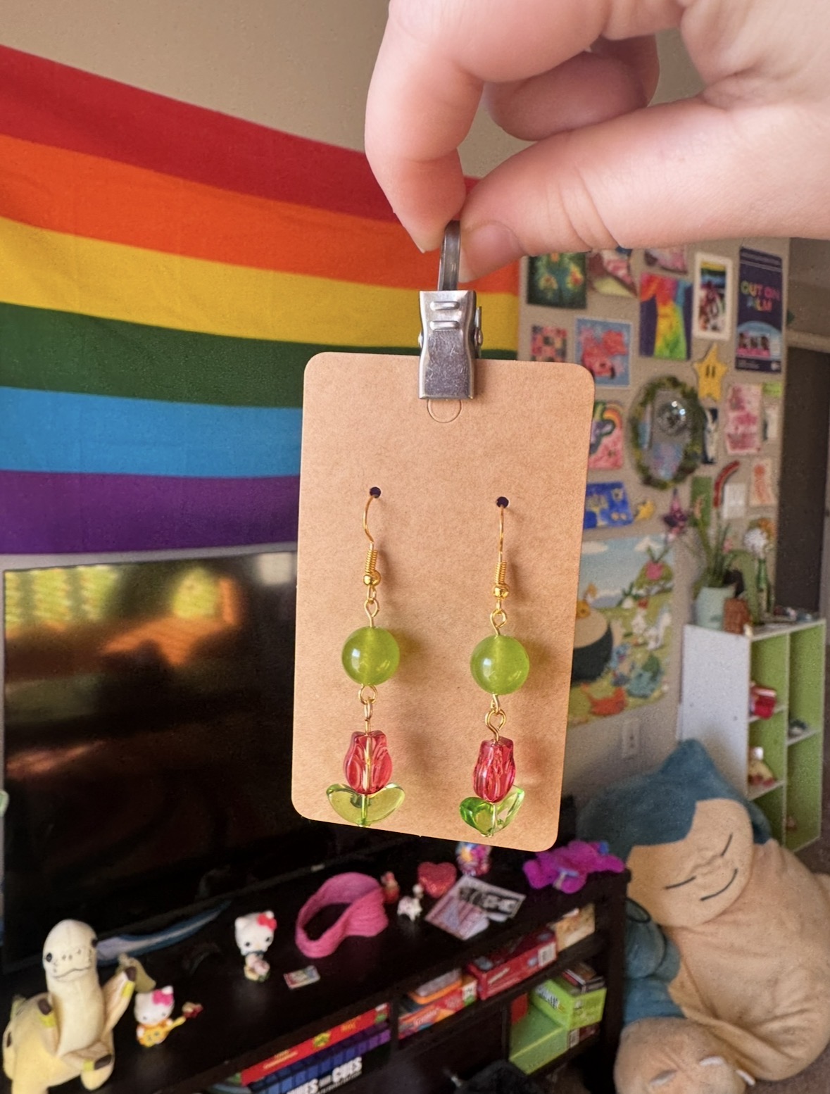 Hand holding a card with green and pink beaded earrings in front of a rainbow flag and a cluttered room.