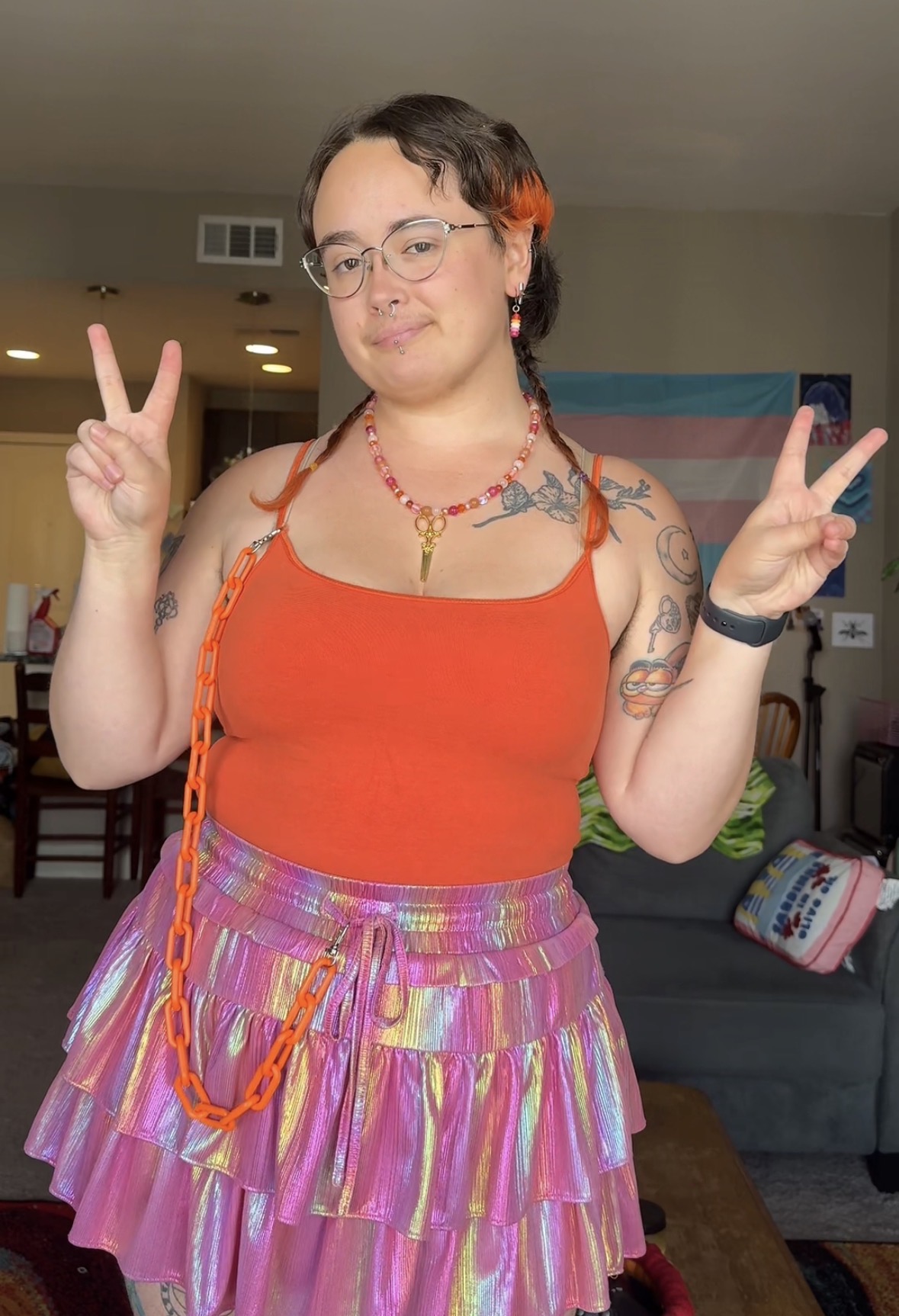 Person with short dark hair, glasses, wearing an orange top and a colorful skirt, making peace signs with both hands.